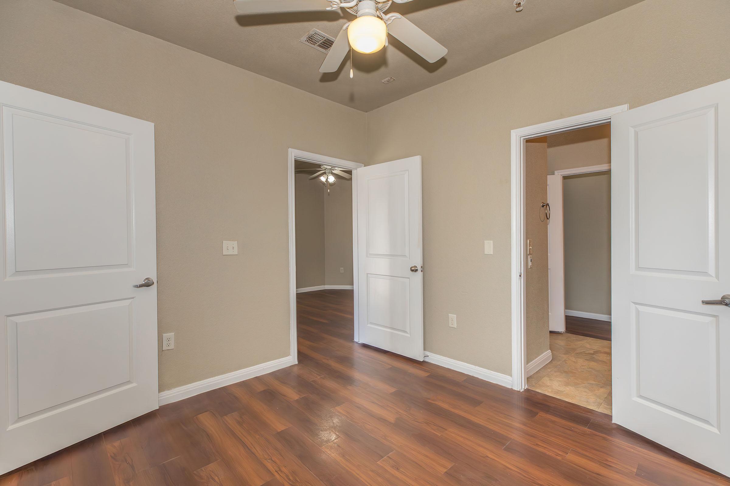 Interior view of a room featuring light brown walls, wooden floors, and multiple doors. One door leads to a closet, and two others open into adjacent spaces. A ceiling fan is visible, adding a modern touch to the cozy atmosphere. The overall space is well-lit and inviting.