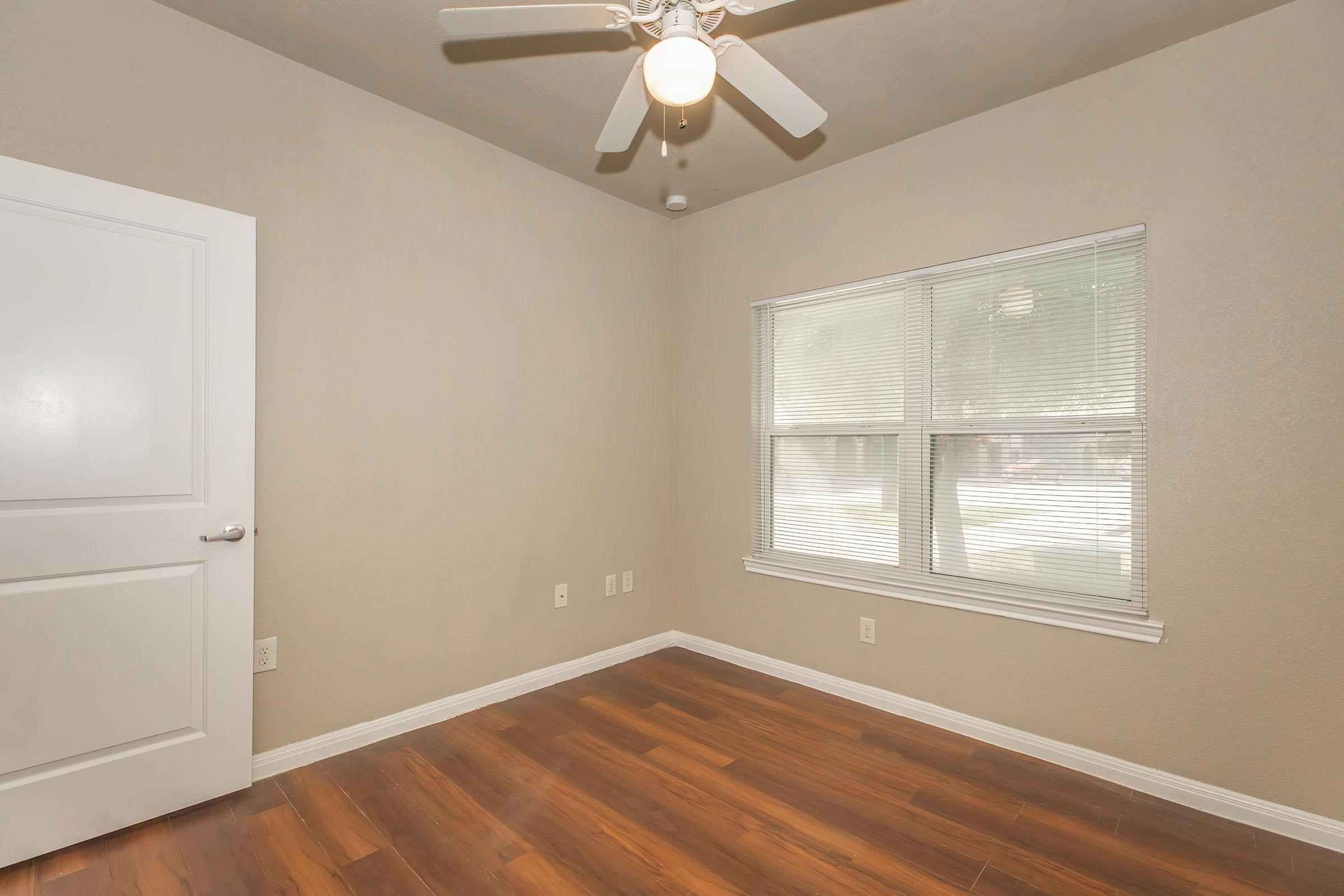 Empty room featuring light brown walls, a ceiling fan with a light fixture, and a window with blinds. The floor is made of dark wood, enhancing the spacious feel of the room. There are electrical outlets on the walls and no furniture present, creating a blank canvas for decorating.