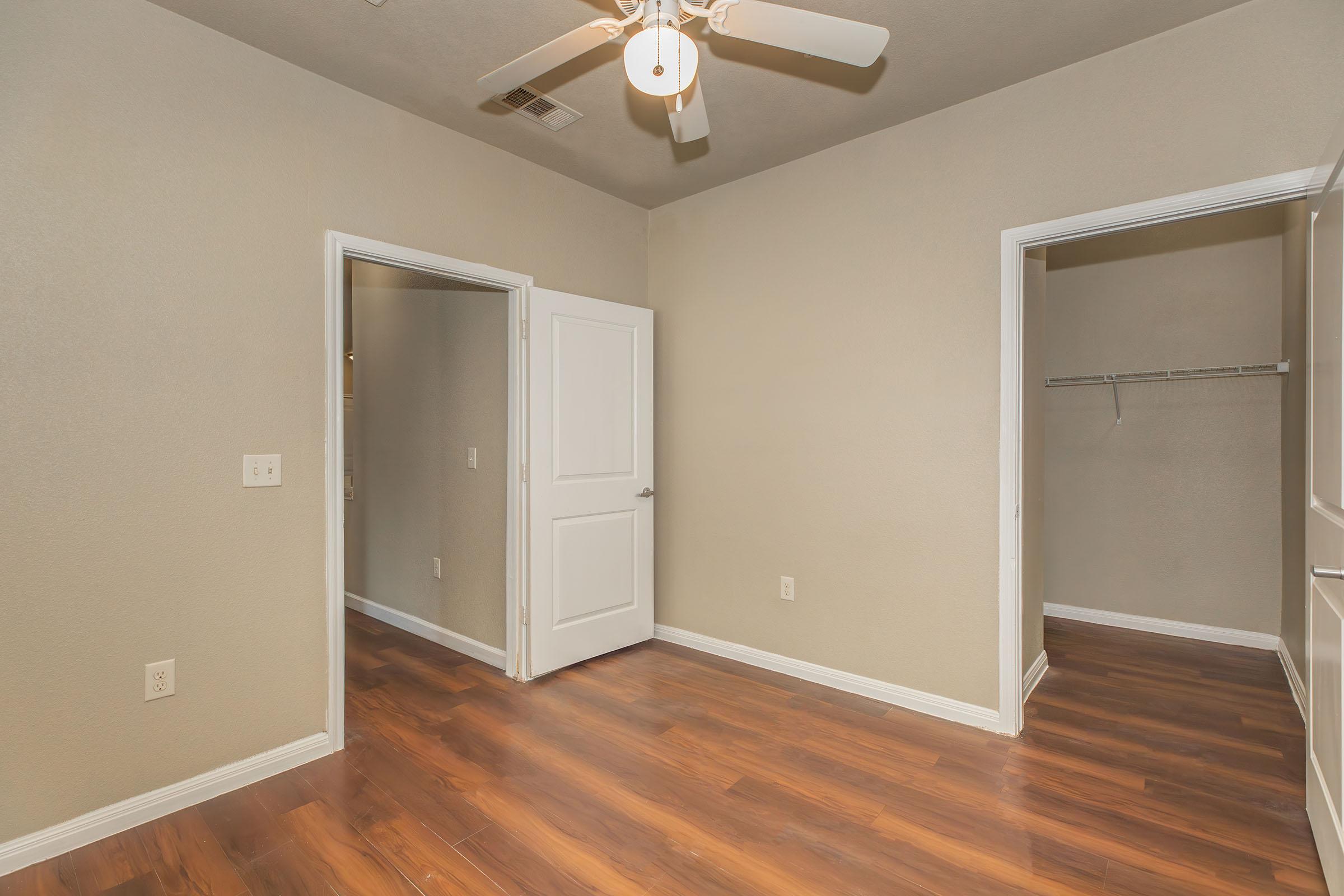 A well-lit room with a ceiling fan, featuring light-colored walls and a hardwood floor. There are two doorways: one leading to another room and the other to a closet with a shelf. The space is empty, creating a clean and minimalist aesthetic.