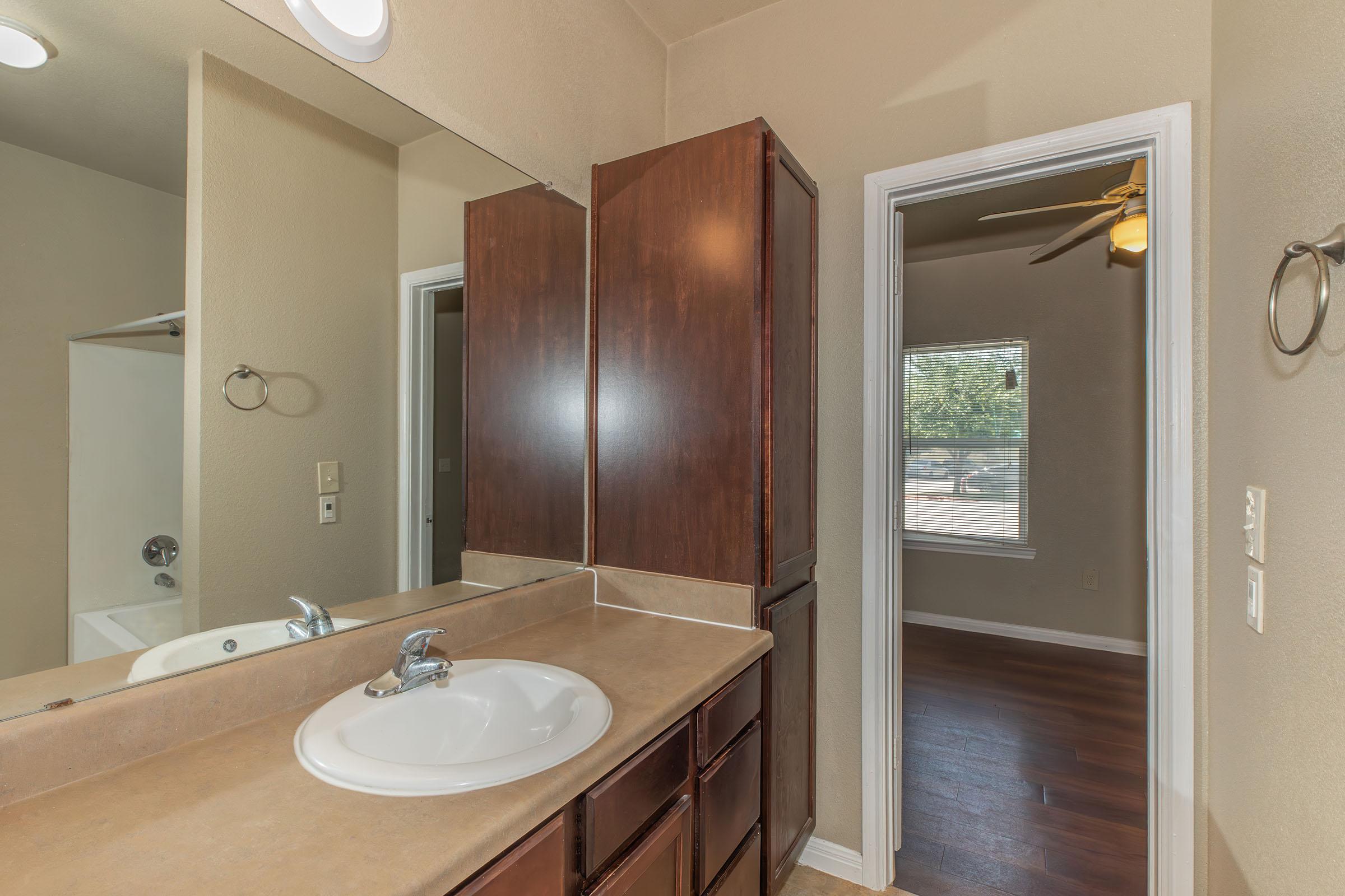 A bathroom featuring a sink with a single faucet, a large mirror on the wall, and wooden cabinets. In the background, an adjoining room is visible through a doorway with a window that lets in natural light. The walls are painted a light color, and the flooring is wooden.