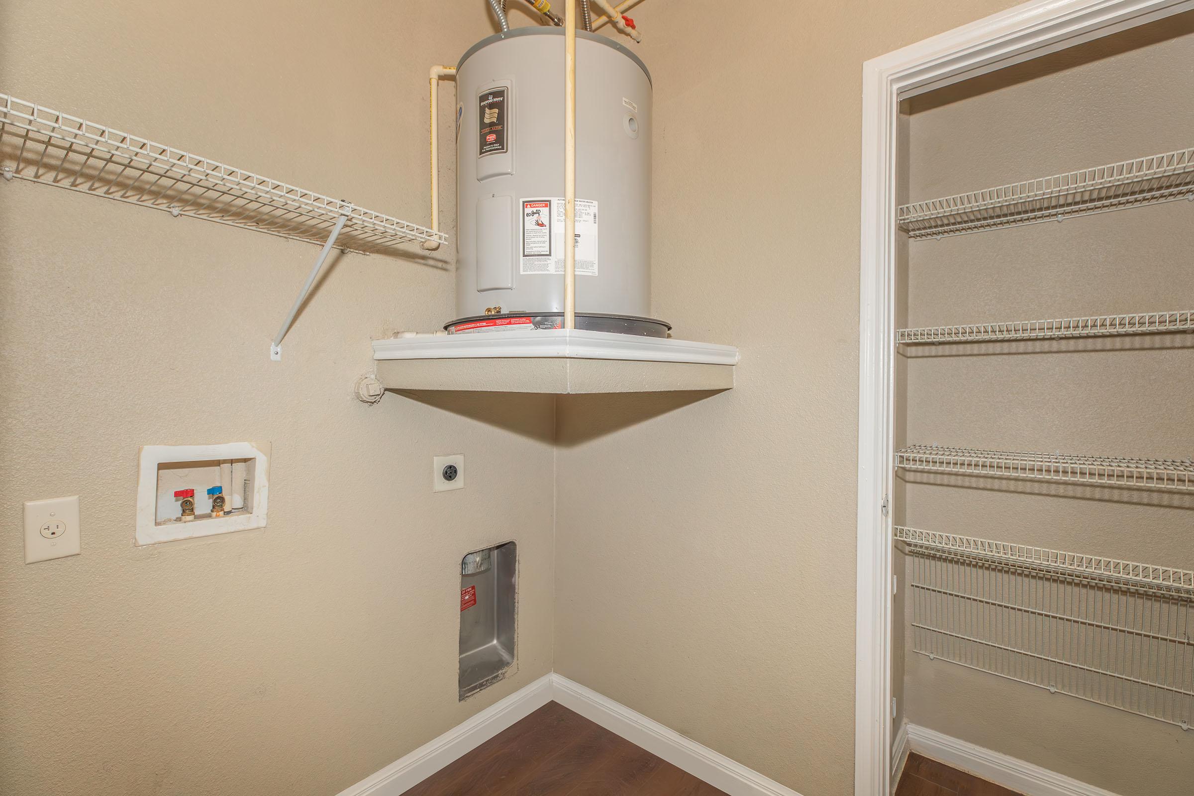 A small utility space featuring a water heater mounted on the wall, with a wire shelf above it. The floor is wooden, and there are empty metal shelves along one wall, providing storage options. The adjacent wall has plumbing fixtures and an electrical outlet. The space has light-colored walls.