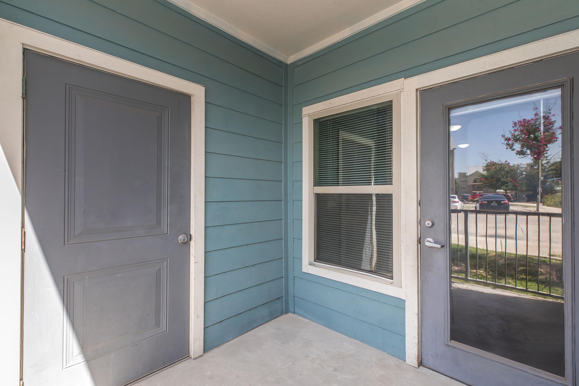 A view of a corner entryway with a grey door on the left and a glass-paneled door on the right. The walls are painted light blue, and a window between the doors offers a glimpse of the outside. Sunlight illuminates the area, highlighting the clean, modern design of the entrance.