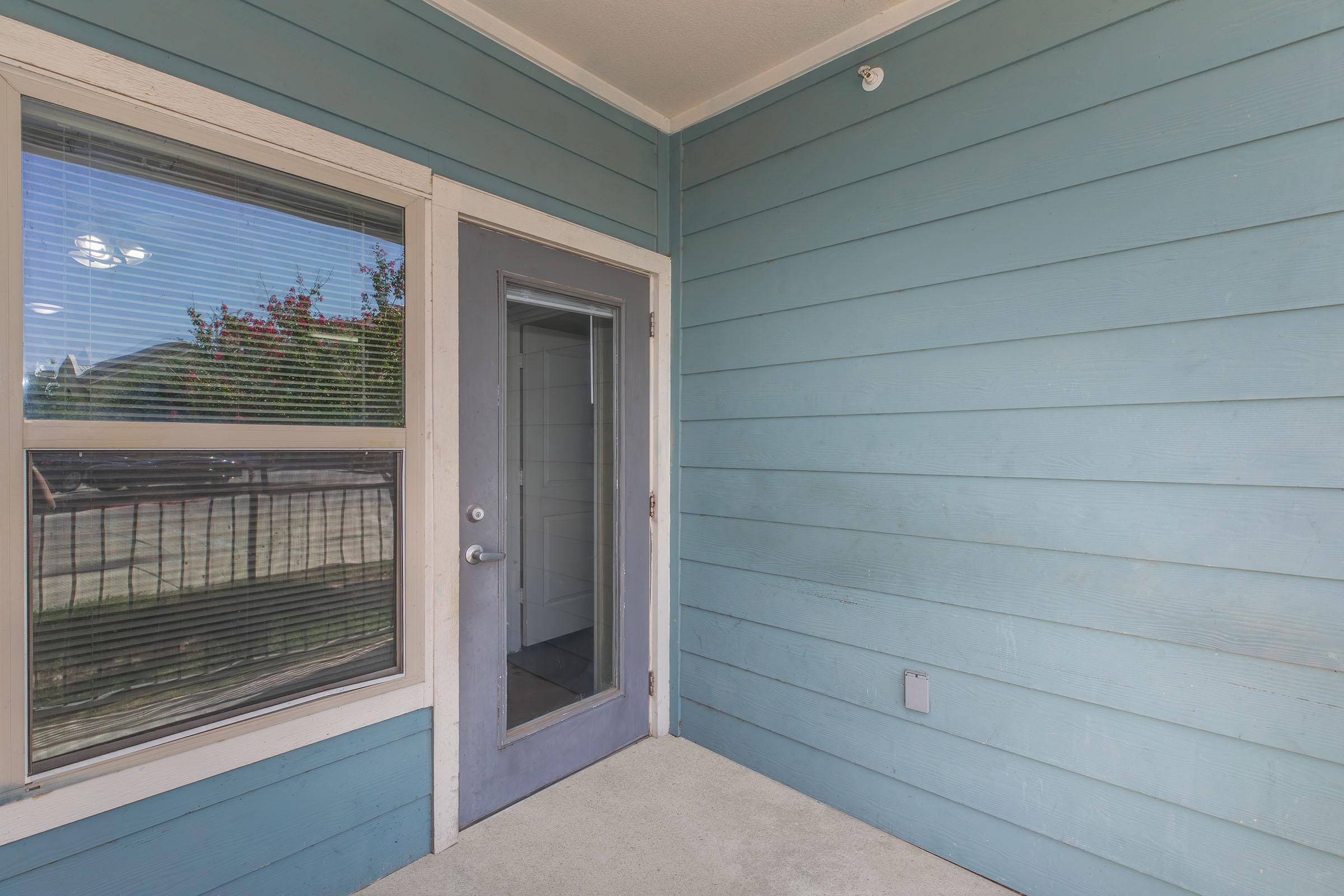 Exterior view of a blue wooden wall featuring a door with a glass panel. To the left, there is a window with horizontal blinds, and outside, you can see green foliage. The area is well-lit, suggesting daytime. The entrance is clean and inviting, suitable for a residential setting.