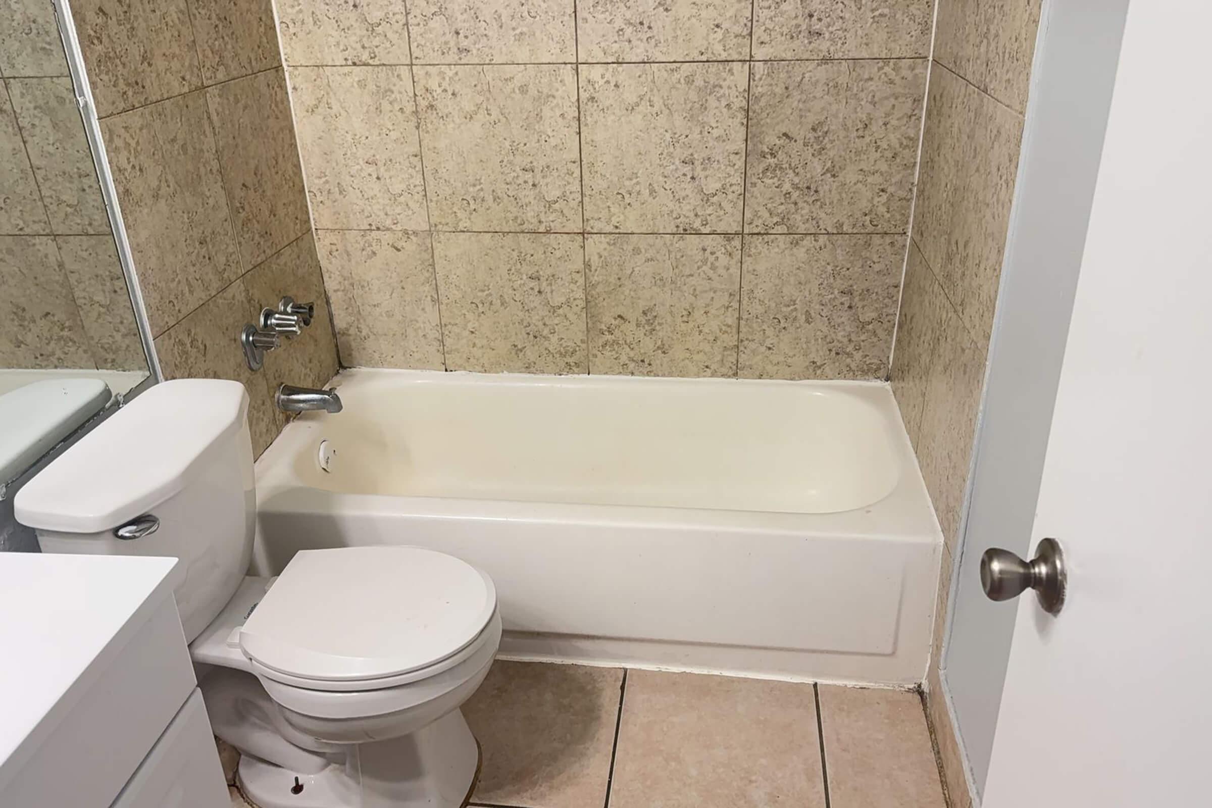 A small bathroom with light-colored tile walls and a beige bathtub. A white toilet is positioned next to the bathtub. There is a mirror above a sink on the left side, with a white cabinet underneath. The floor is tiled, and a white door is visible on the right side of the image.