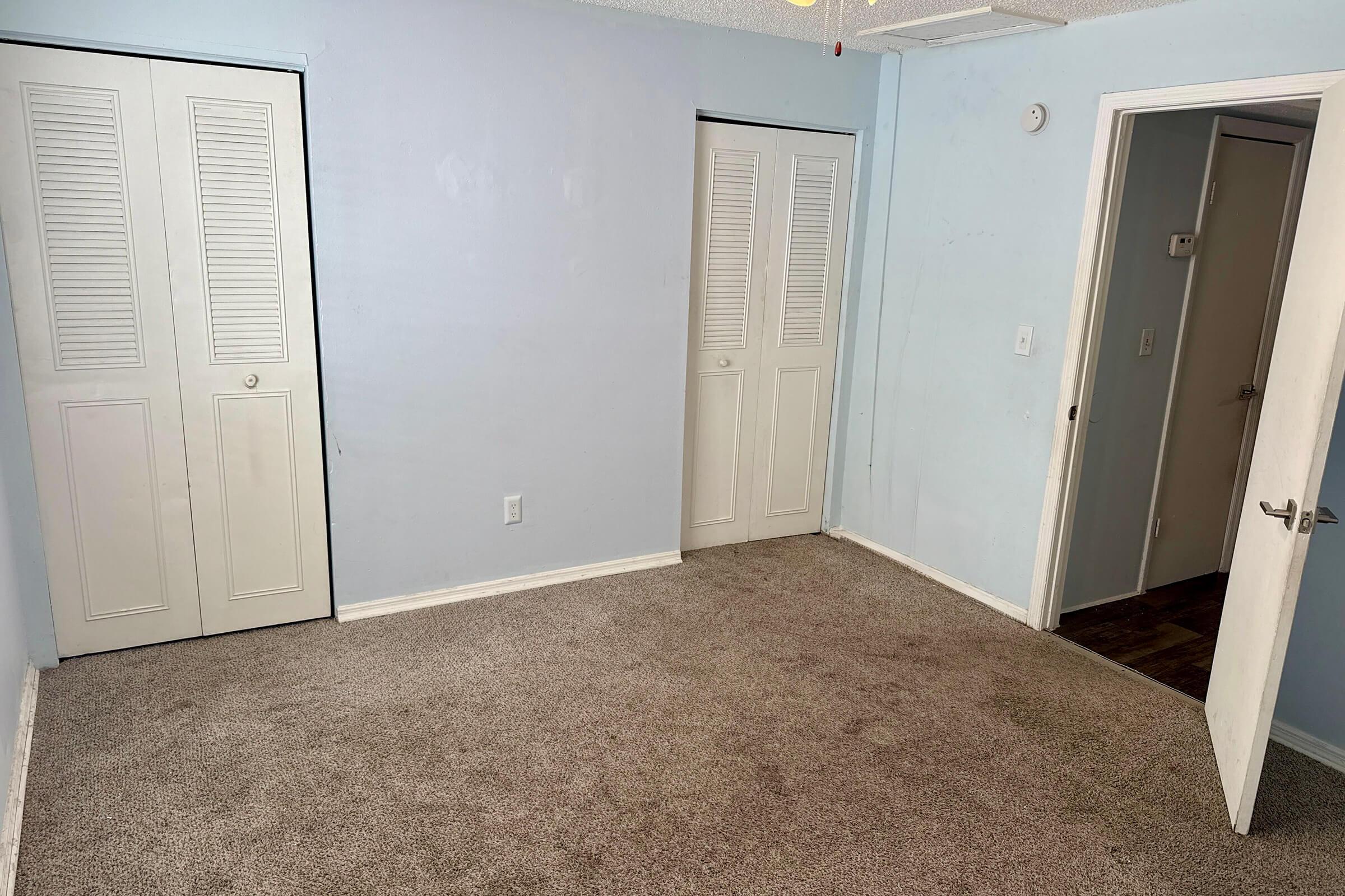 A vacant room with light blue walls and beige carpet. There are two white closet doors on the left and a door on the right leading to a hallway. Natural light is coming from the doorway, creating a simple and clean atmosphere in the space.