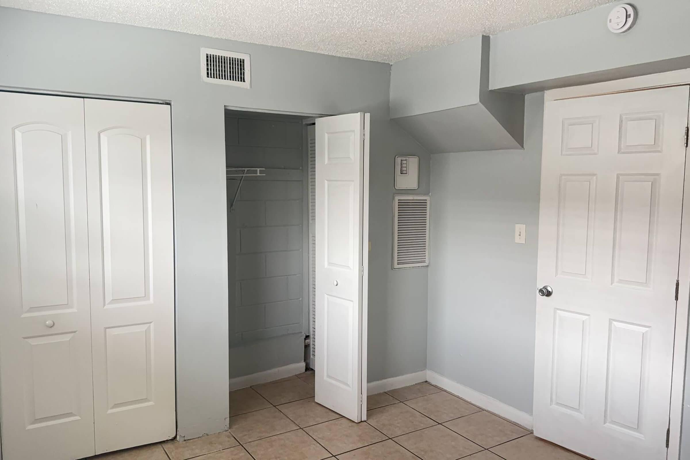 A small room with light gray walls and tiled flooring. There are two white closet doors on the left, an open door leading into a closet on the right, and a third door further along the wall. An air vent and a small wall-mounted unit are visible. The space is well-lit and uncluttered.