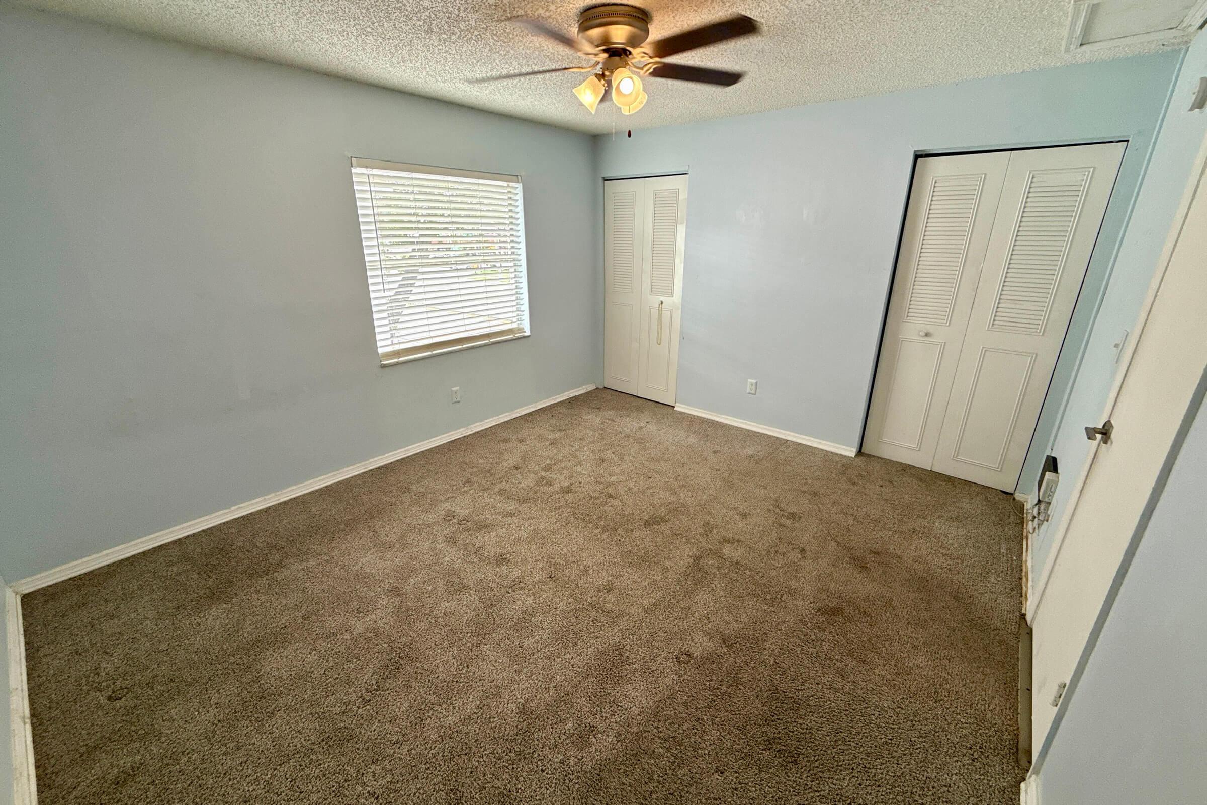 A well-lit, empty bedroom featuring a ceiling fan, light blue walls, and beige carpet. There is a window with blinds on one side and two closed closet doors on the opposite wall, creating a spacious and clean appearance.
