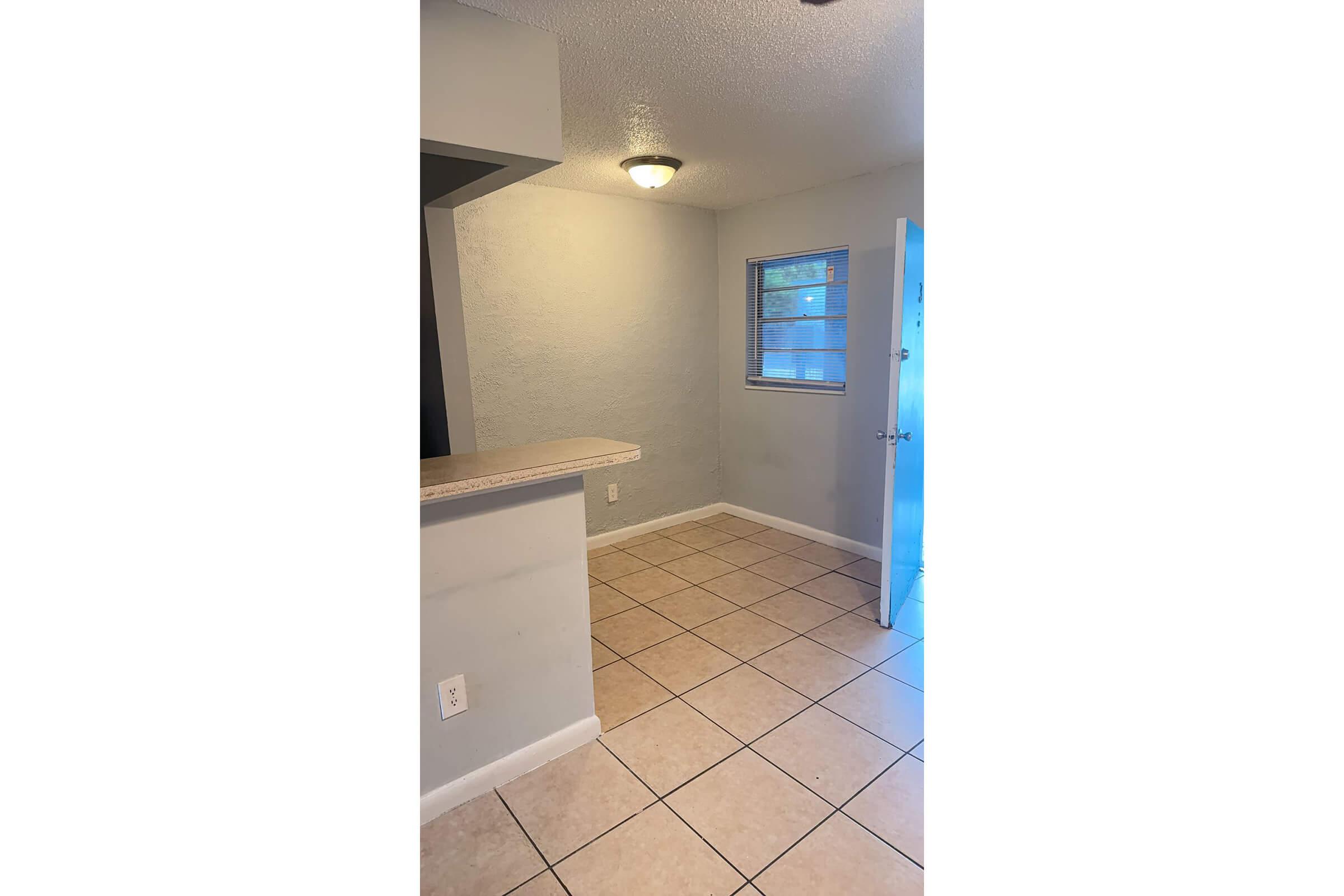 A small room with light gray walls and beige tiled flooring. There's a doorway on the right leading outside, a window on the wall ahead, and a counter on the left. The ceiling light provides illumination, creating a bright and open feel.
