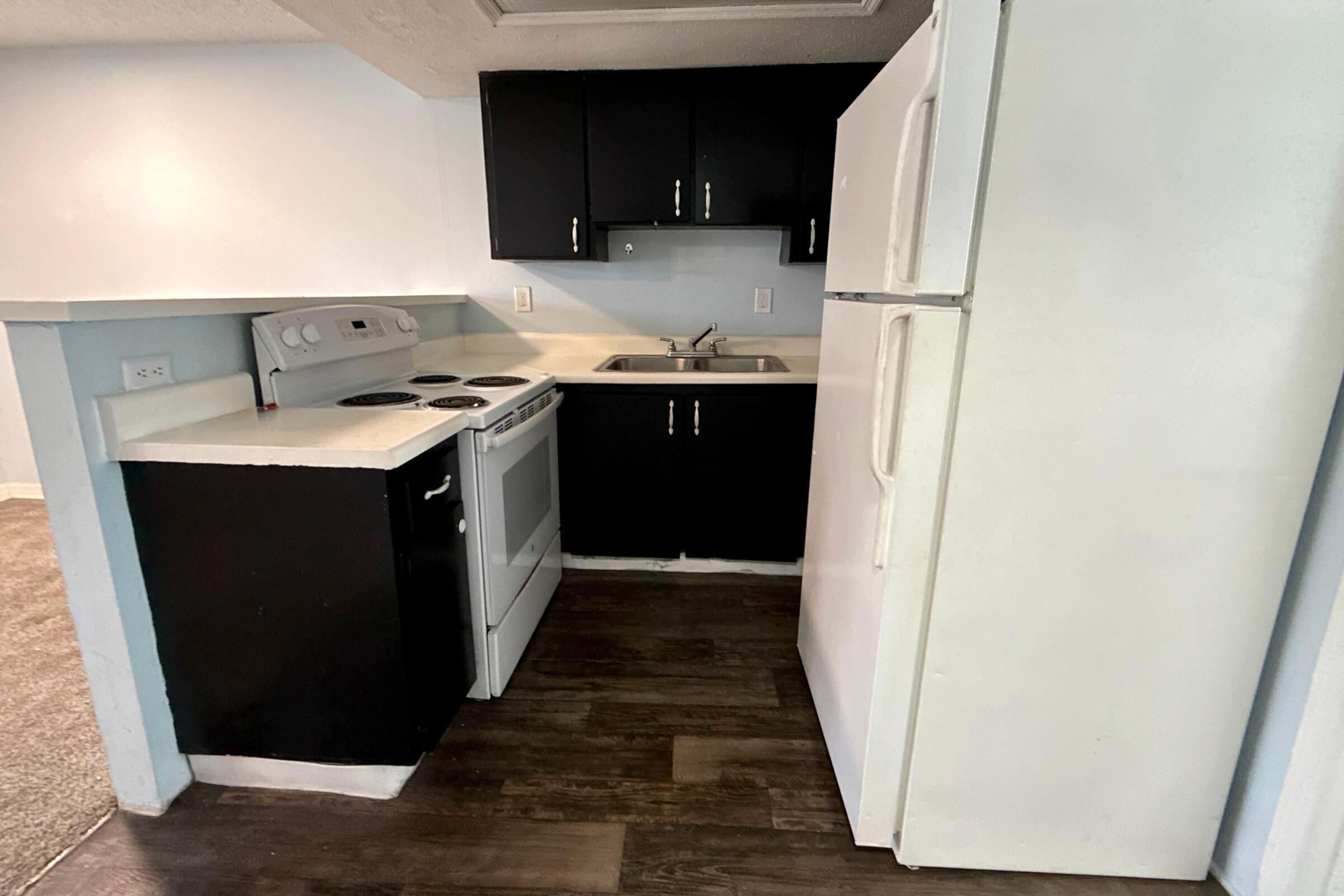 A compact kitchen featuring a black cabinet with a white countertop, a white refrigerator, and a white electric stove with an oven. The walls are light blue, and the flooring is dark wood. There is a sink with a faucet, and the kitchen appears well-lit and organized.