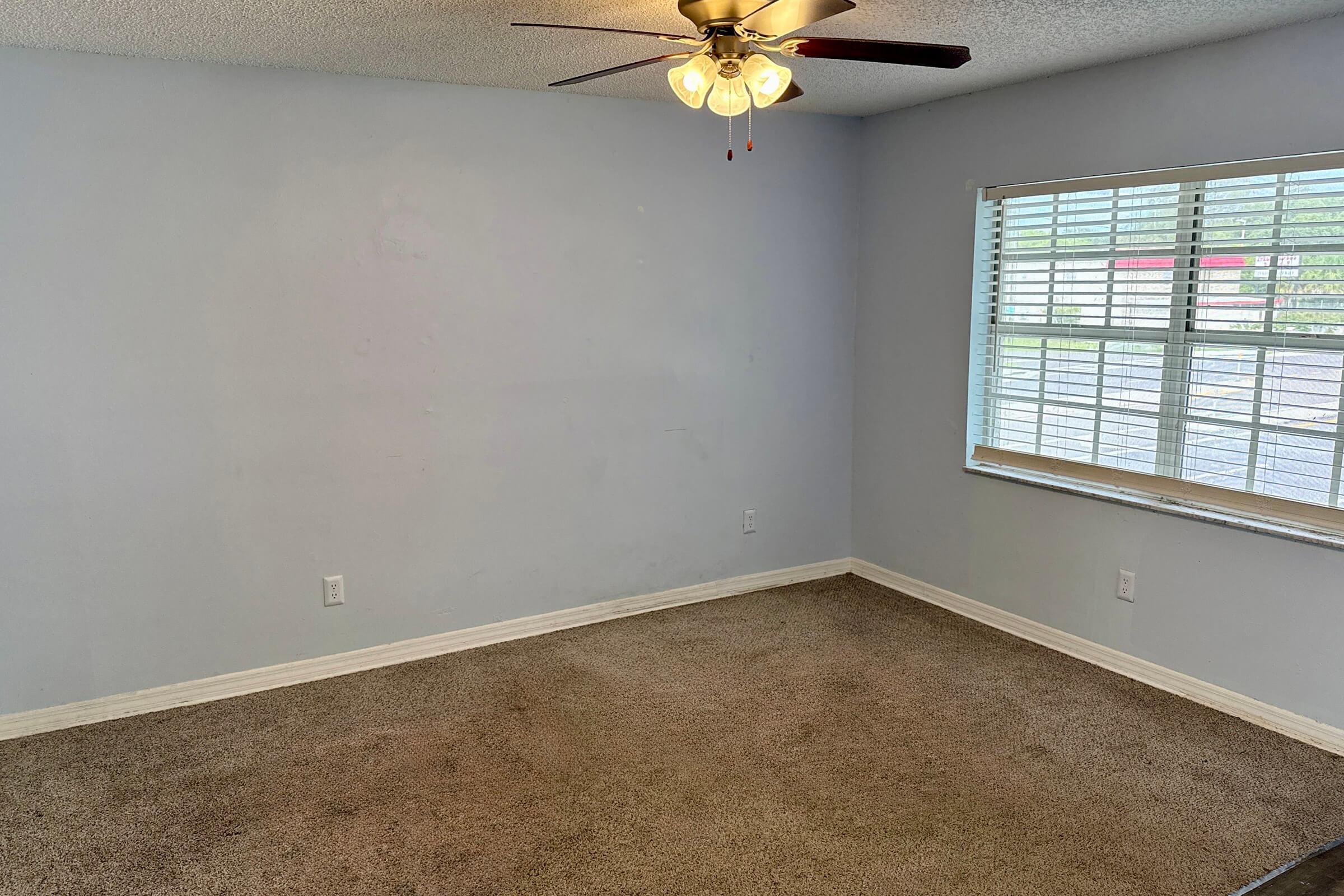 Empty room with light purple walls, carpeted floor, and a ceiling fan. A window with white blinds allows natural light in. The space appears clean and uncluttered, suggesting a neutral setting suitable for various uses.