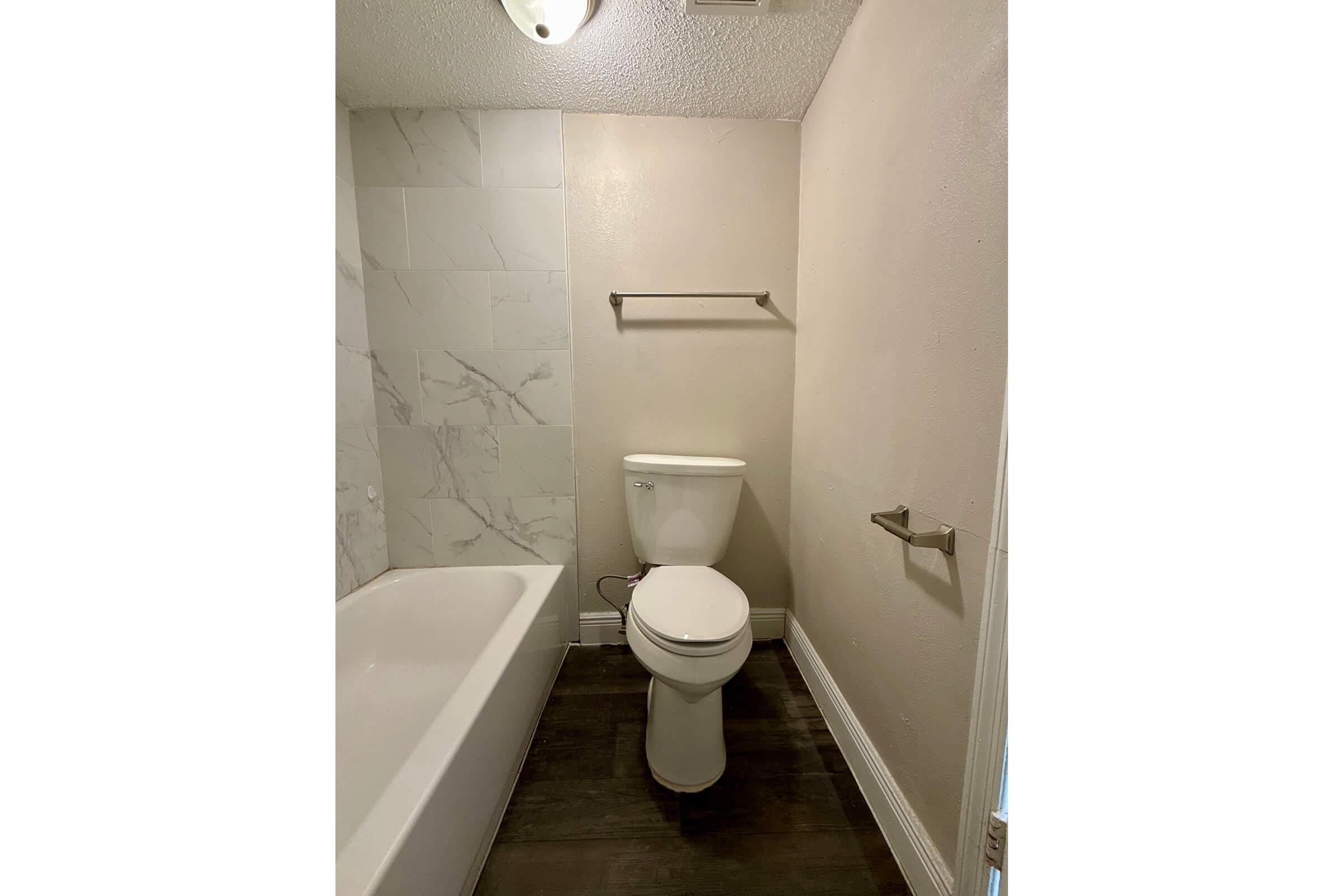 A small bathroom featuring a white toilet and bathtub. The walls are painted a light beige, with one side showcasing patterned white tiles. A simple shelf is mounted above the toilet, and there is a towel bar next to it. A light fixture is visible on the ceiling. The floor has dark wood-like tiles.