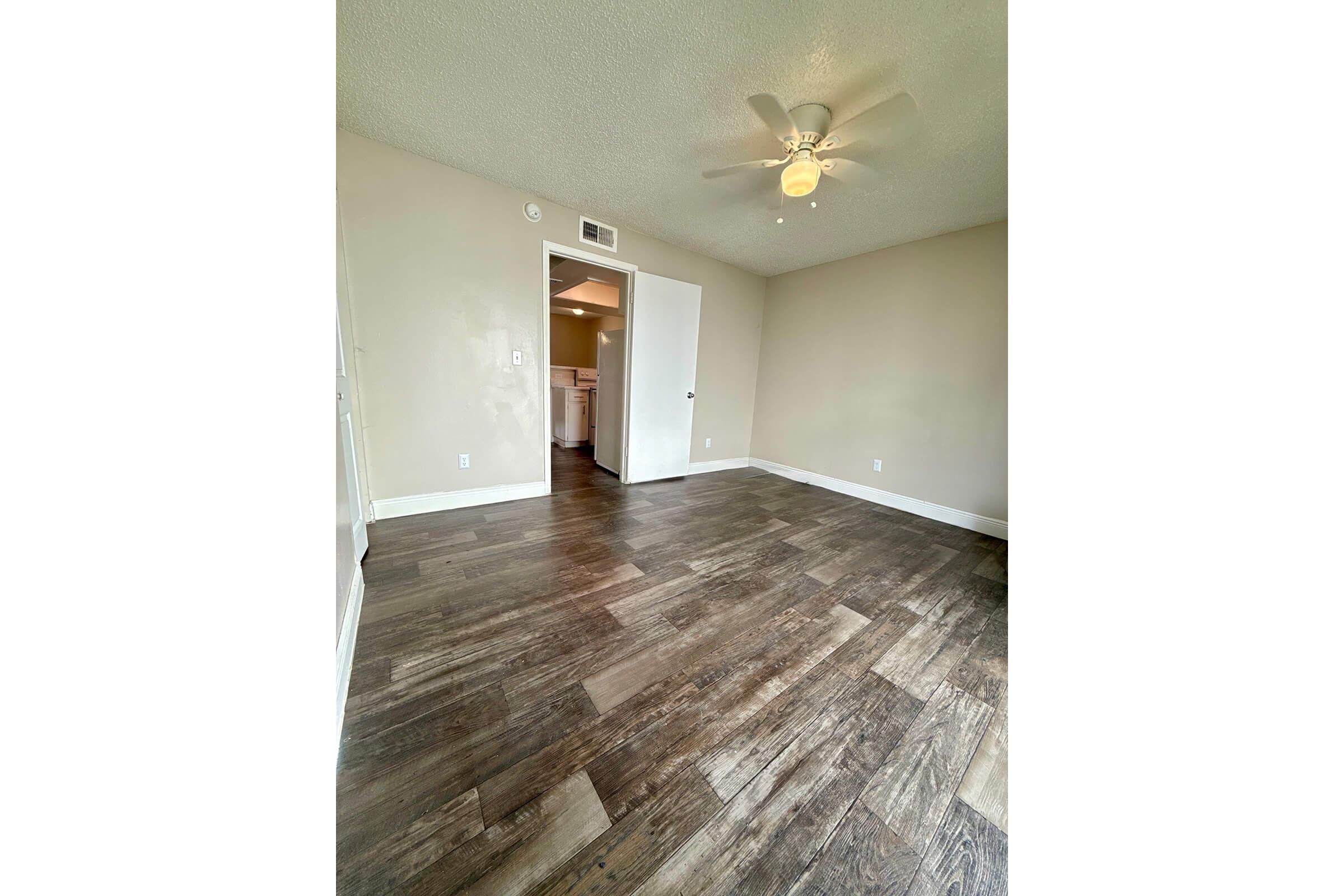 Empty room with light brown wooden flooring, a ceiling fan, and a single door leading to another room. The walls are painted a light beige color. Natural light enters from an unseen window, creating a bright and spacious feel.