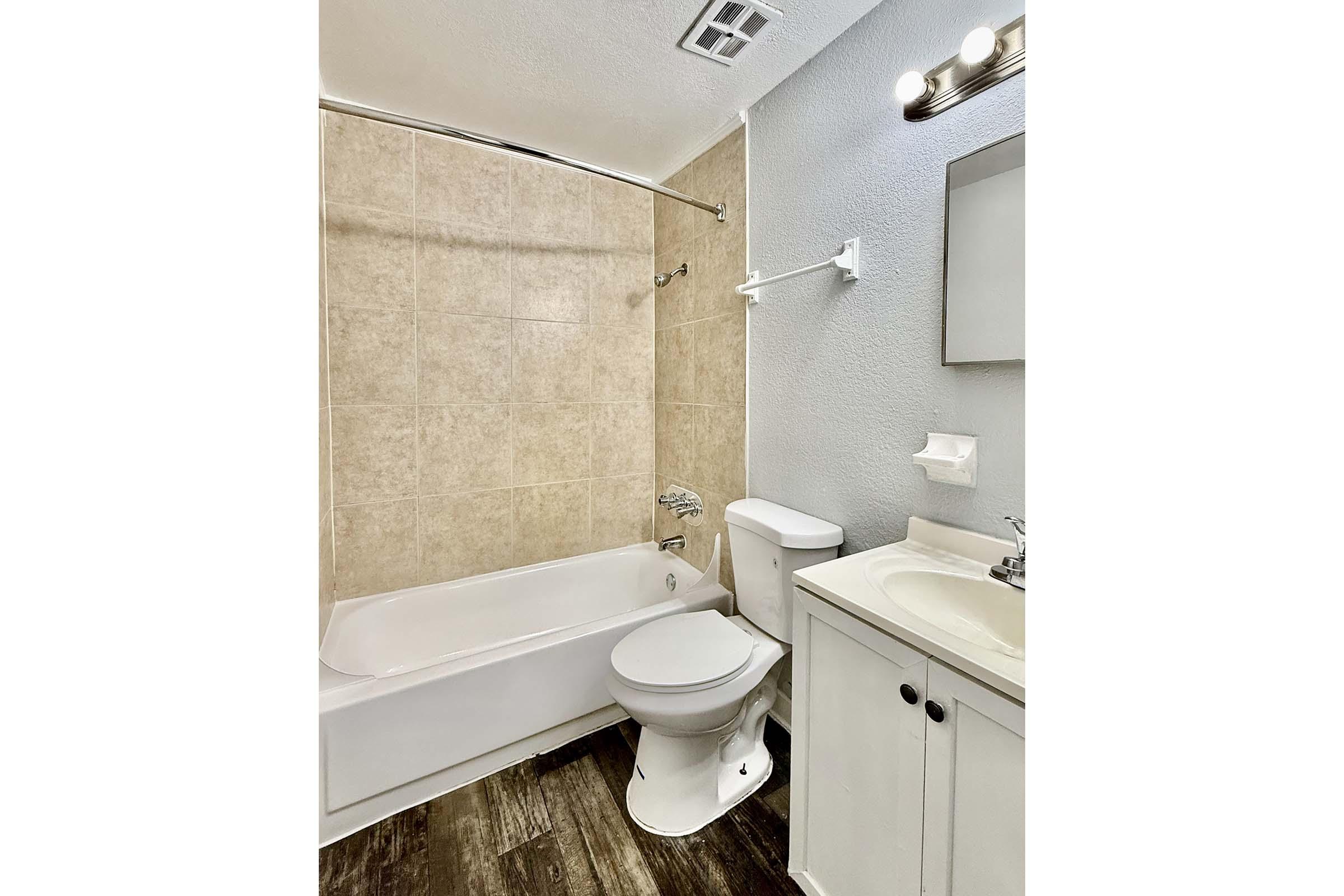 A clean and modern bathroom featuring a white bathtub and shower combination, a toilet, and a small vanity with a sink. The walls are a soft blue, and the floor is dark wood. There is a mirror above the sink and a towel rack on the wall. Natural light illuminates the space.