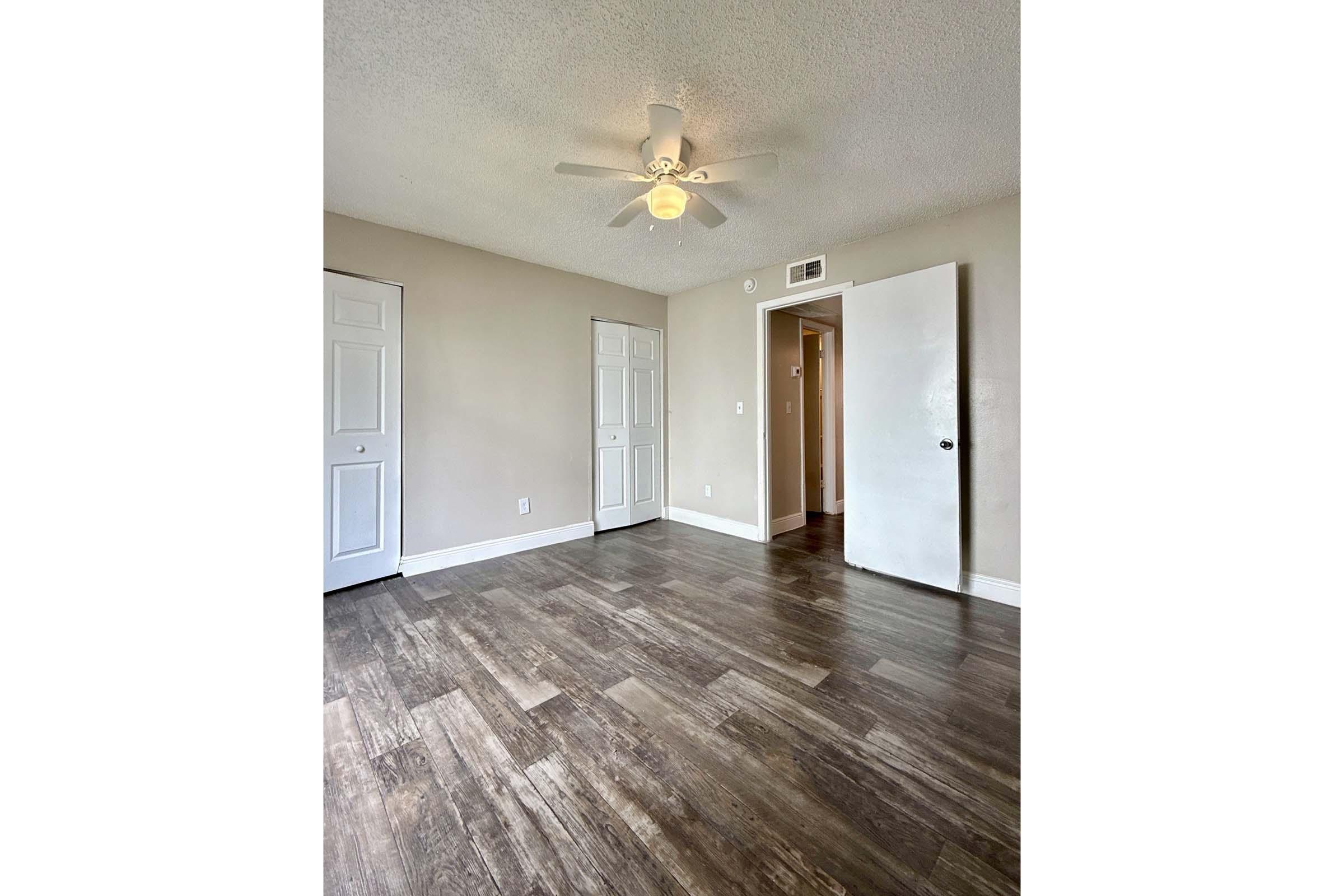 A spacious room with light-colored walls and a ceiling fan. The floor is finished with wood-like laminate. There are two white doors on the left side and a single white door on the right, leading to a closet. Natural light is coming through an unseen window, creating a bright atmosphere.