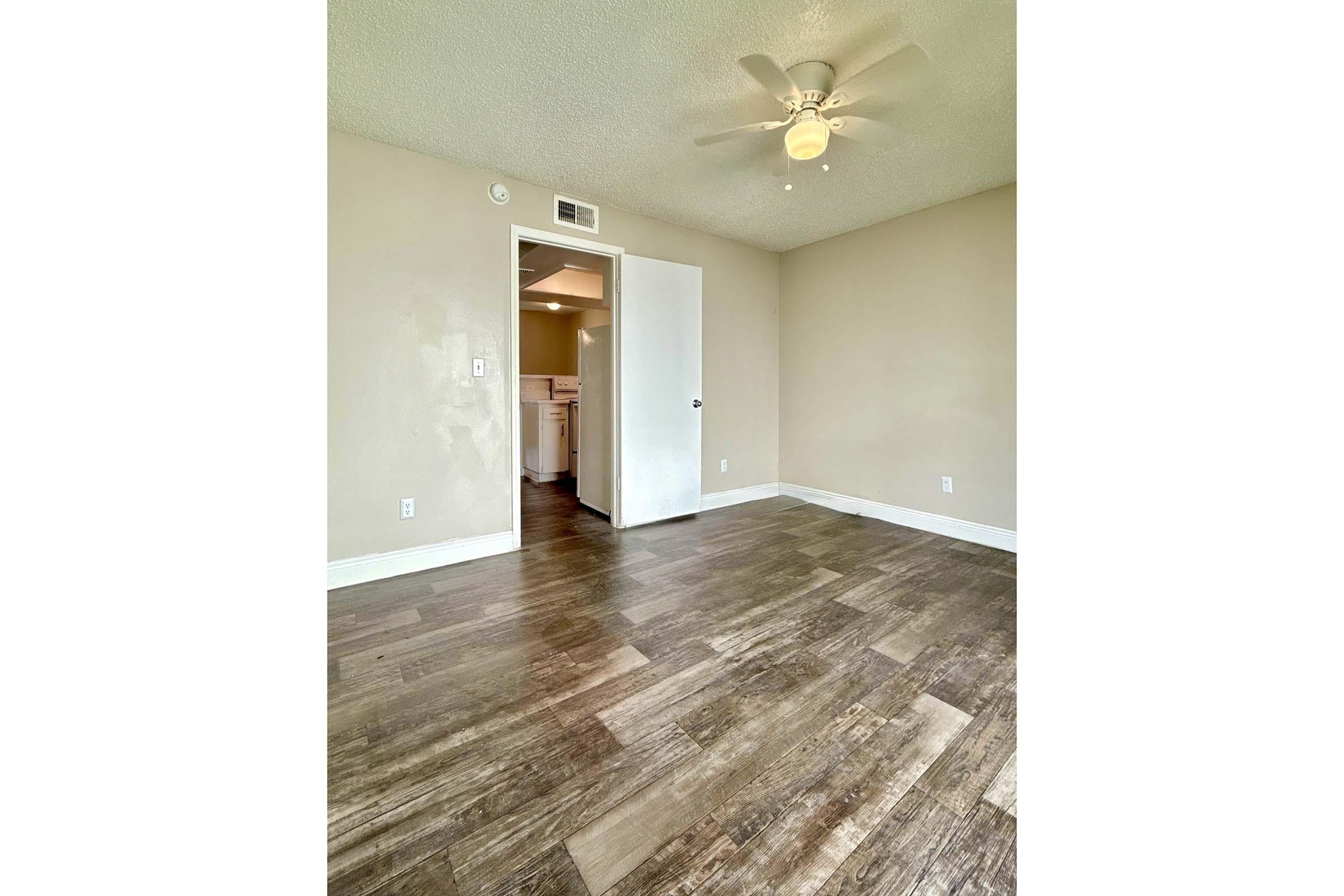 A spacious room with light brown wood-like flooring, a ceiling fan, and a light beige wall. A closed white door leads to a kitchen area with visible light and cabinetry. The room is well-lit, creating an inviting atmosphere.