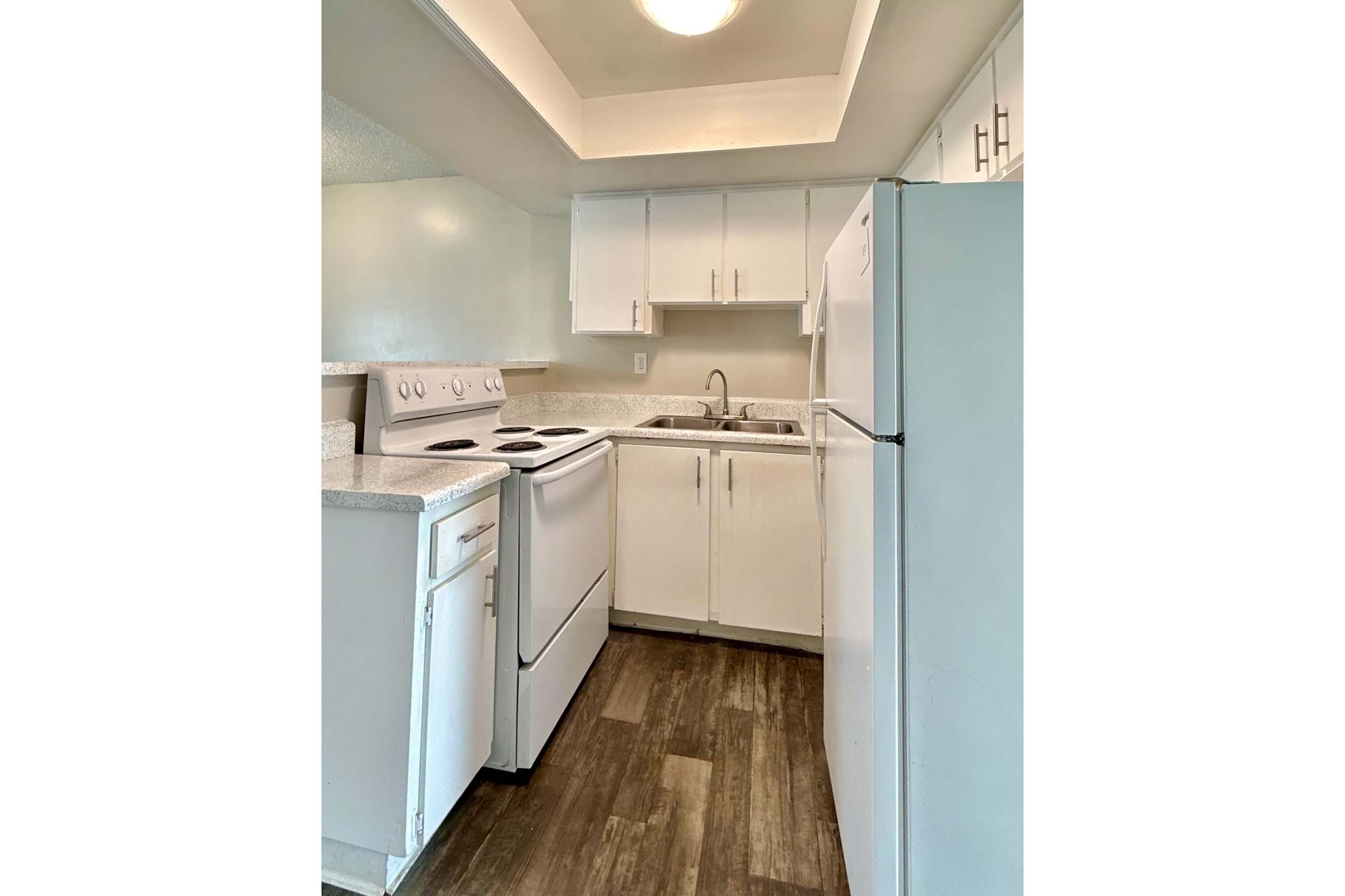 A modern kitchen featuring white cabinetry, a countertop, a double sink, and white appliances including an oven, stove, and refrigerator. The flooring is dark wood, providing a contrast to the light-colored walls and cabinets. Natural light is coming from a ceiling fixture.