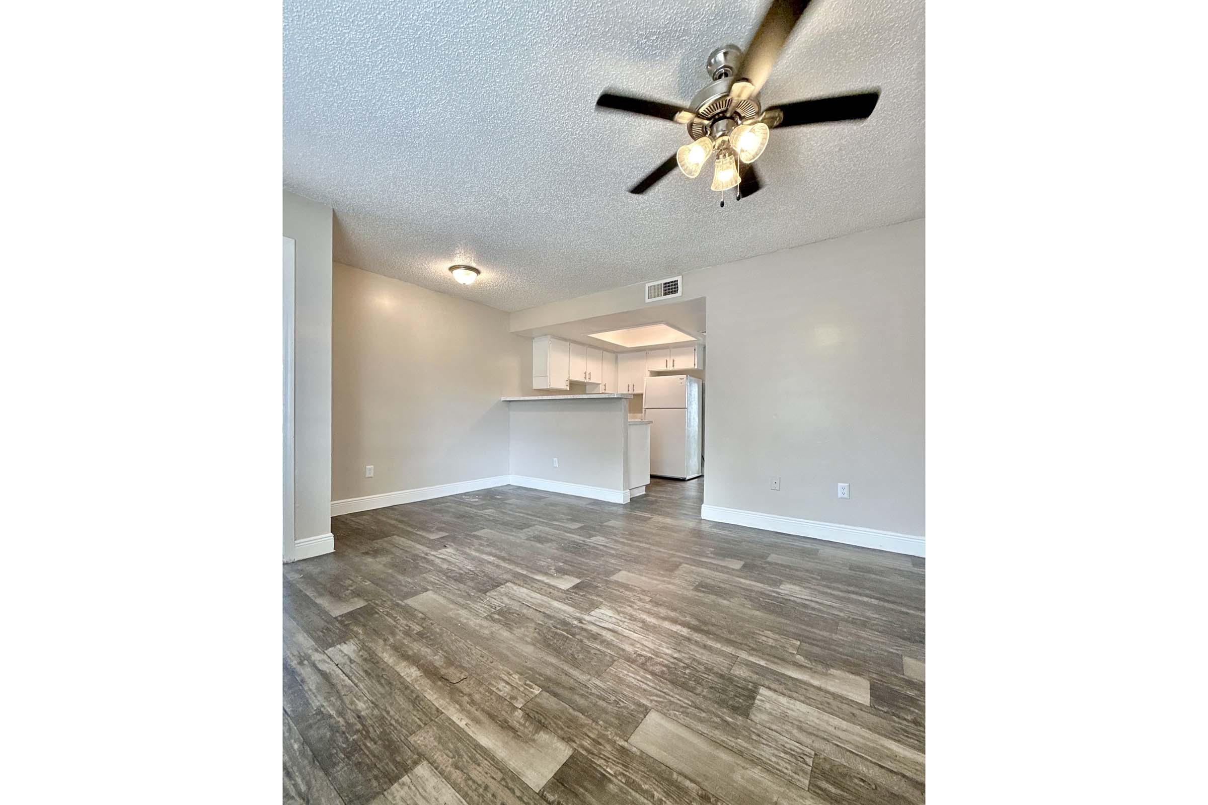 A spacious living area featuring light-colored walls and a ceiling fan. The floor is covered with stylish wood-like tiles. In the background, a kitchen is visible, showcasing white cabinetry and appliances. Natural light brightens the room, creating a welcoming atmosphere.