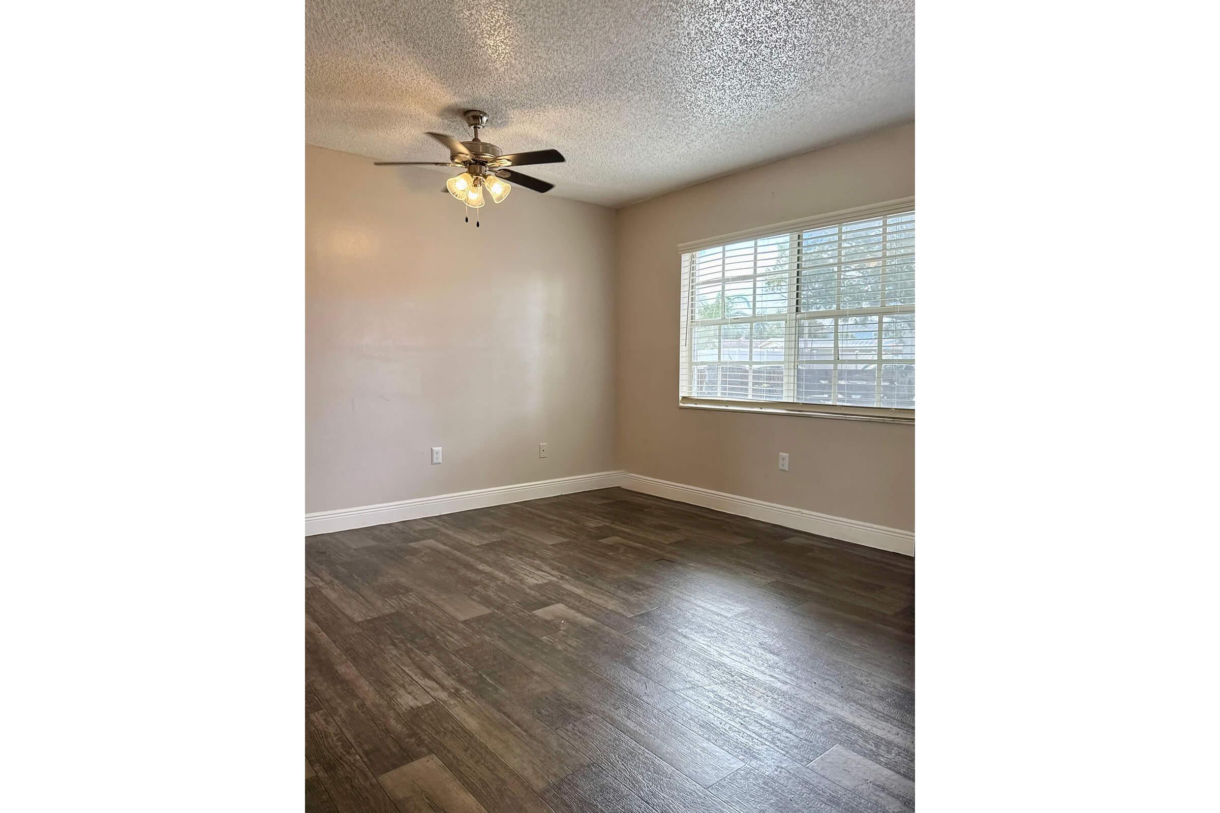 A clean, empty room with a light-colored wall, a ceiling fan with four light bulbs, and a window with white blinds. The floor features brown, wooden-like tiles, creating a spacious and well-lit ambiance.