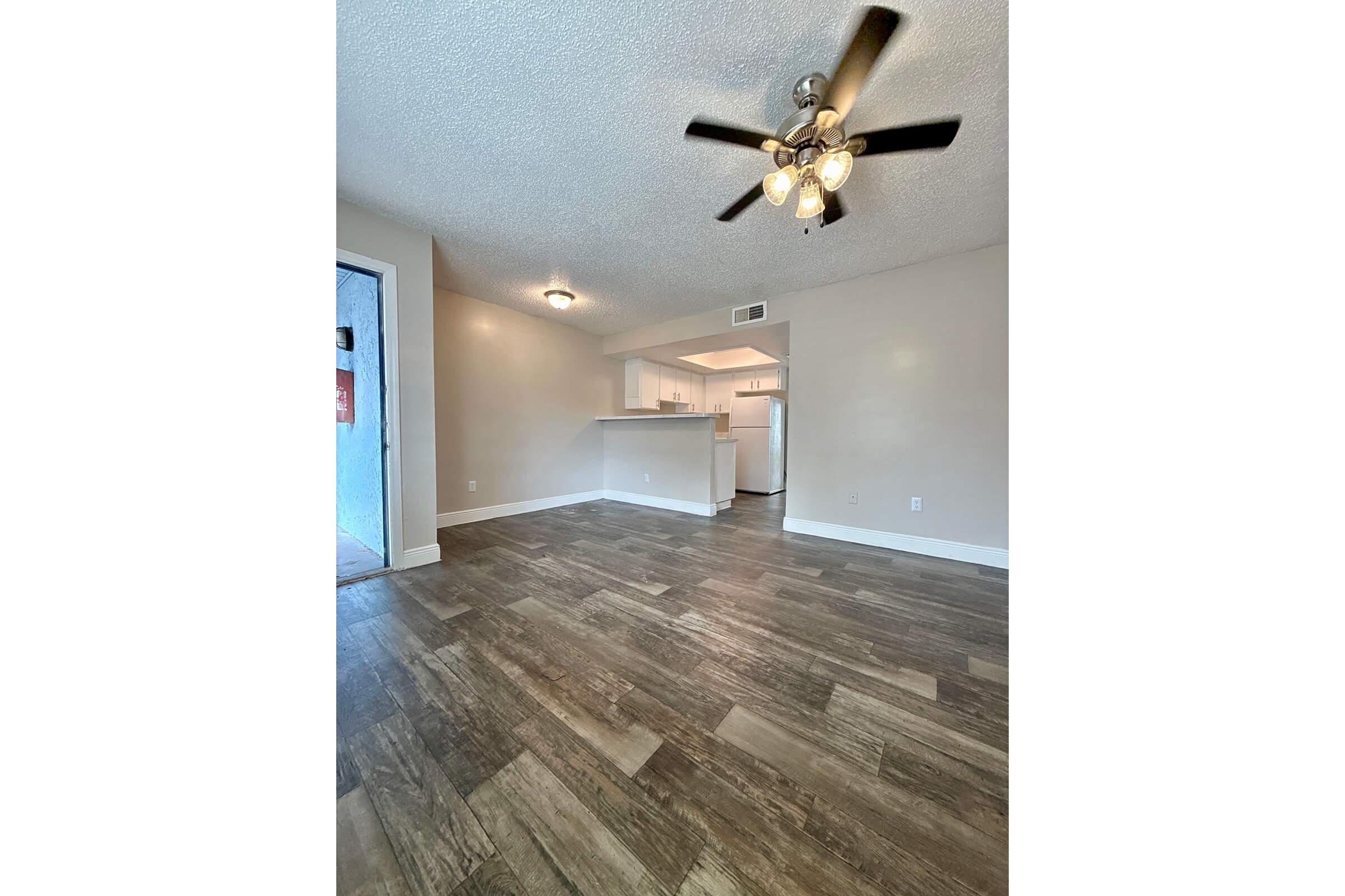 Spacious living area featuring a light-colored ceiling with a ceiling fan, modern laminate flooring, and neutral walls. The open layout includes a view of a kitchen with white appliances and cabinetry in the background. Natural light enters from a nearby window, creating a bright atmosphere.