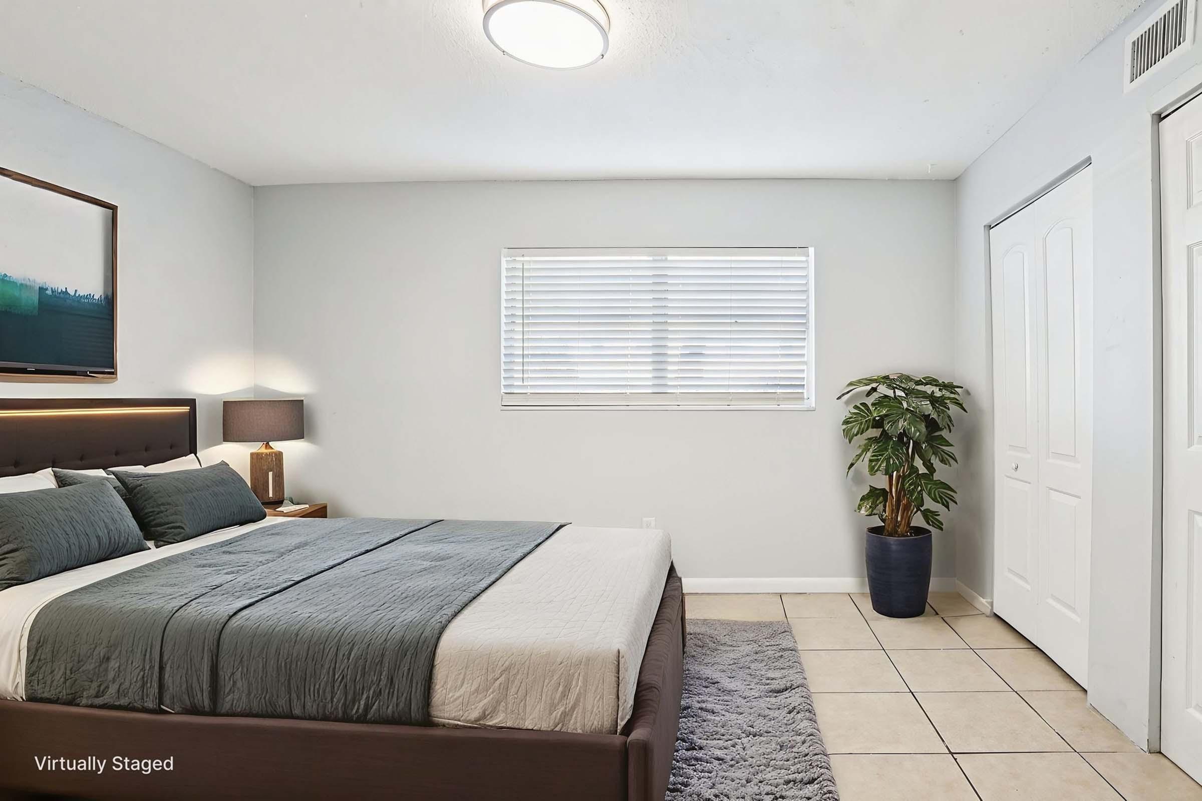 A cozy bedroom featuring a large bed with gray linens, lamps on either side, a potted plant in the corner, and a window with blinds. The walls are painted light gray, and the floor is tiled. A piece of art hangs above the bed, adding a touch of color to the space.