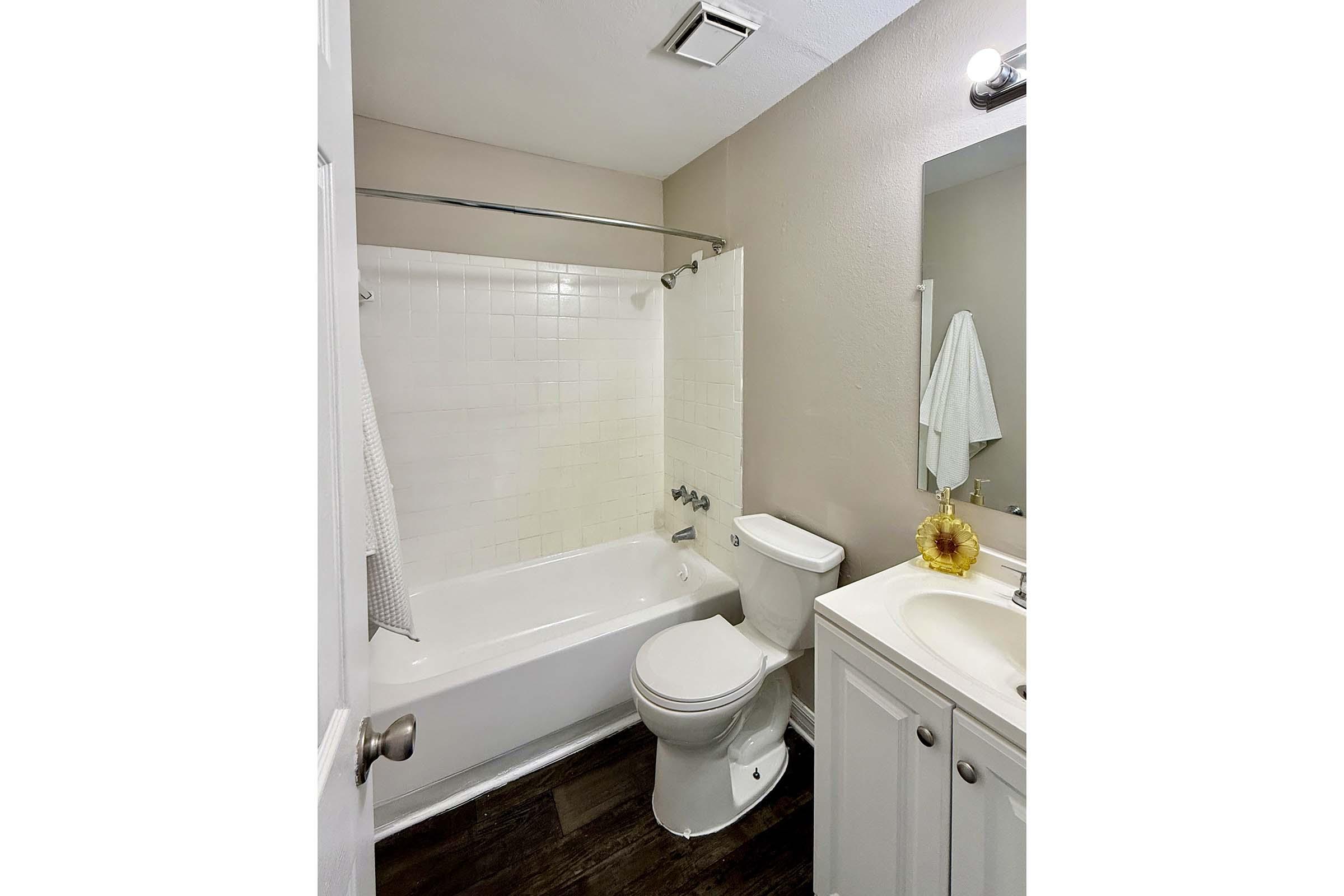 A clean and modern bathroom featuring a white bathtub and shower, a toilet, and a single sink vanity with a mirror. The walls are painted light, and there is a towel hanging on a rack. A sunflower decoration adds a touch of color. The flooring is dark wood, contrasting with the lighter elements.