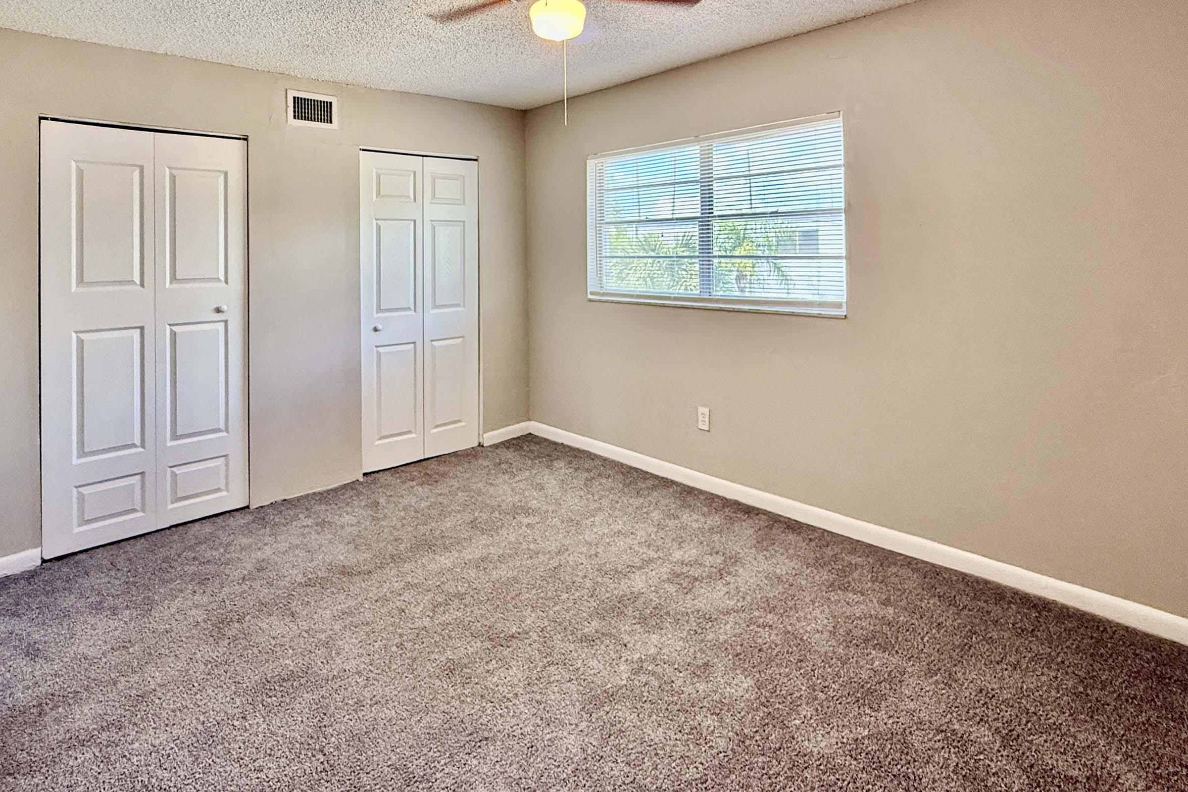 A clean, empty room with light grey carpet, two closed white doors on the left, and a window with white blinds letting in natural light. The walls are painted a soft beige, and a ceiling fan is visible near the center of the room.