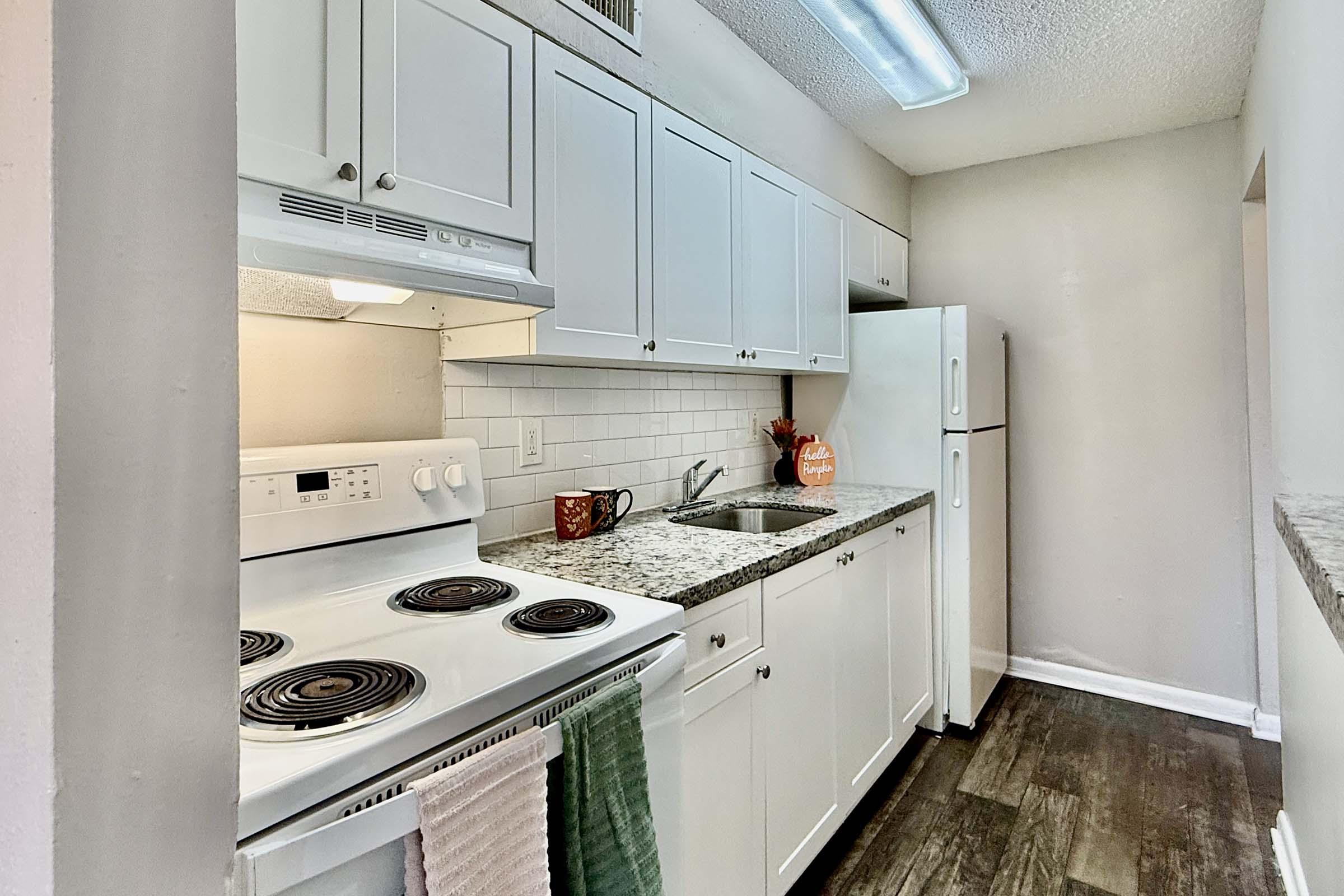 A modern kitchen featuring white cabinetry, a white oven and refrigerator, and a granite countertop with a sink. A small plant and utensils are placed on the counter, and there are light-colored walls and flooring that enhance the space's bright and airy feel.