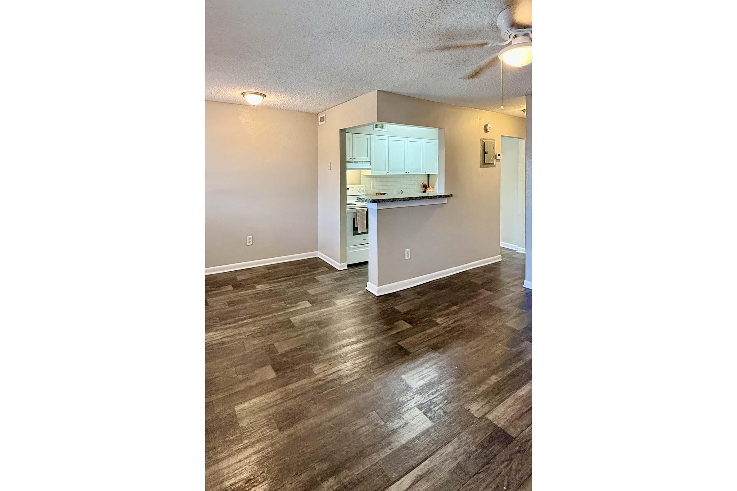A spacious, well-lit room featuring wood-like flooring, a ceiling fan, and light-colored walls. In the background, there is a kitchen area with white cabinets and appliances visible. The layout suggests an open concept living space, creating a welcoming atmosphere.