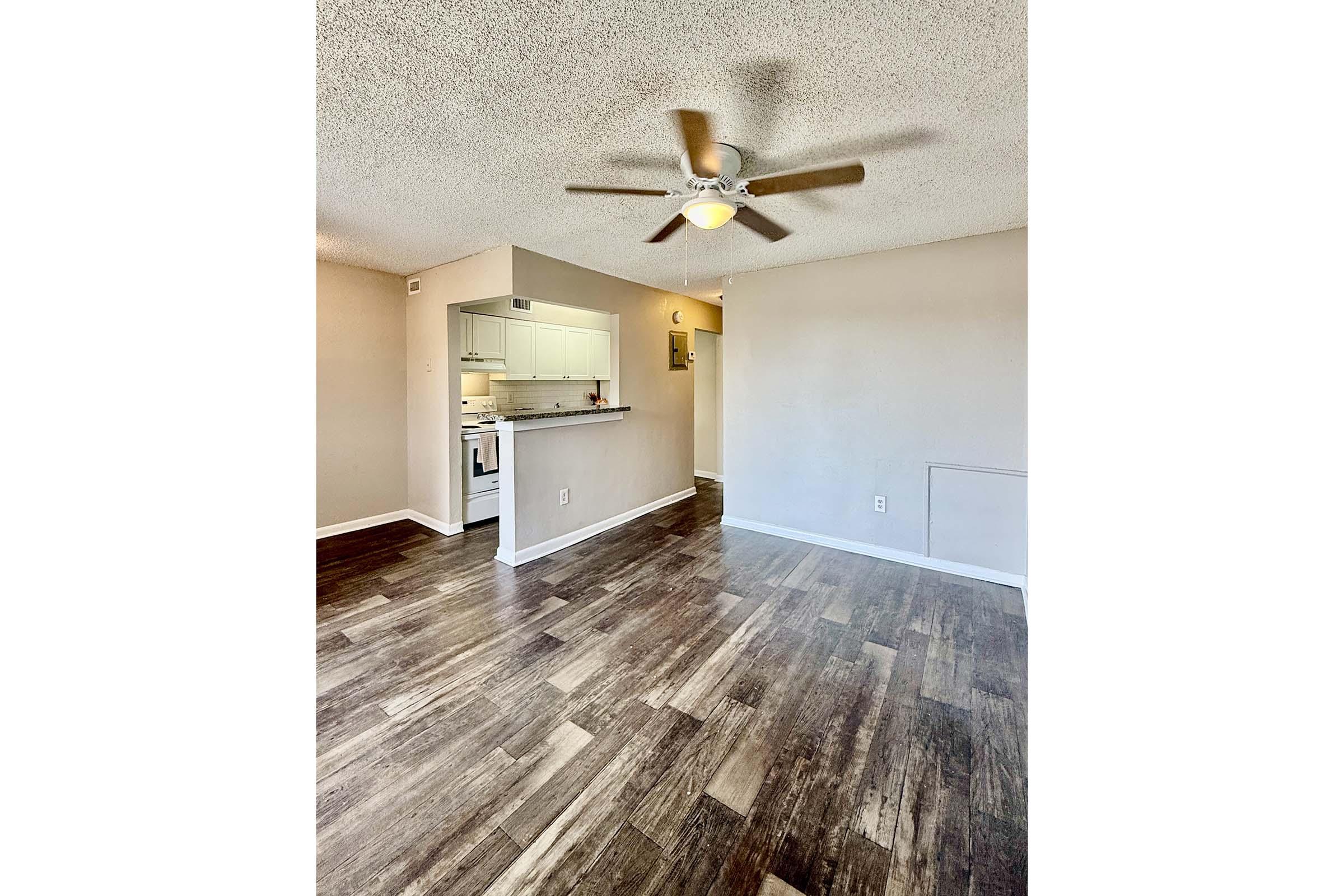 A spacious living area with a ceiling fan, showcasing wood-like flooring. On the left, a glimpse of a kitchen with white cabinetry. The walls are painted light colors, creating a warm and inviting atmosphere.