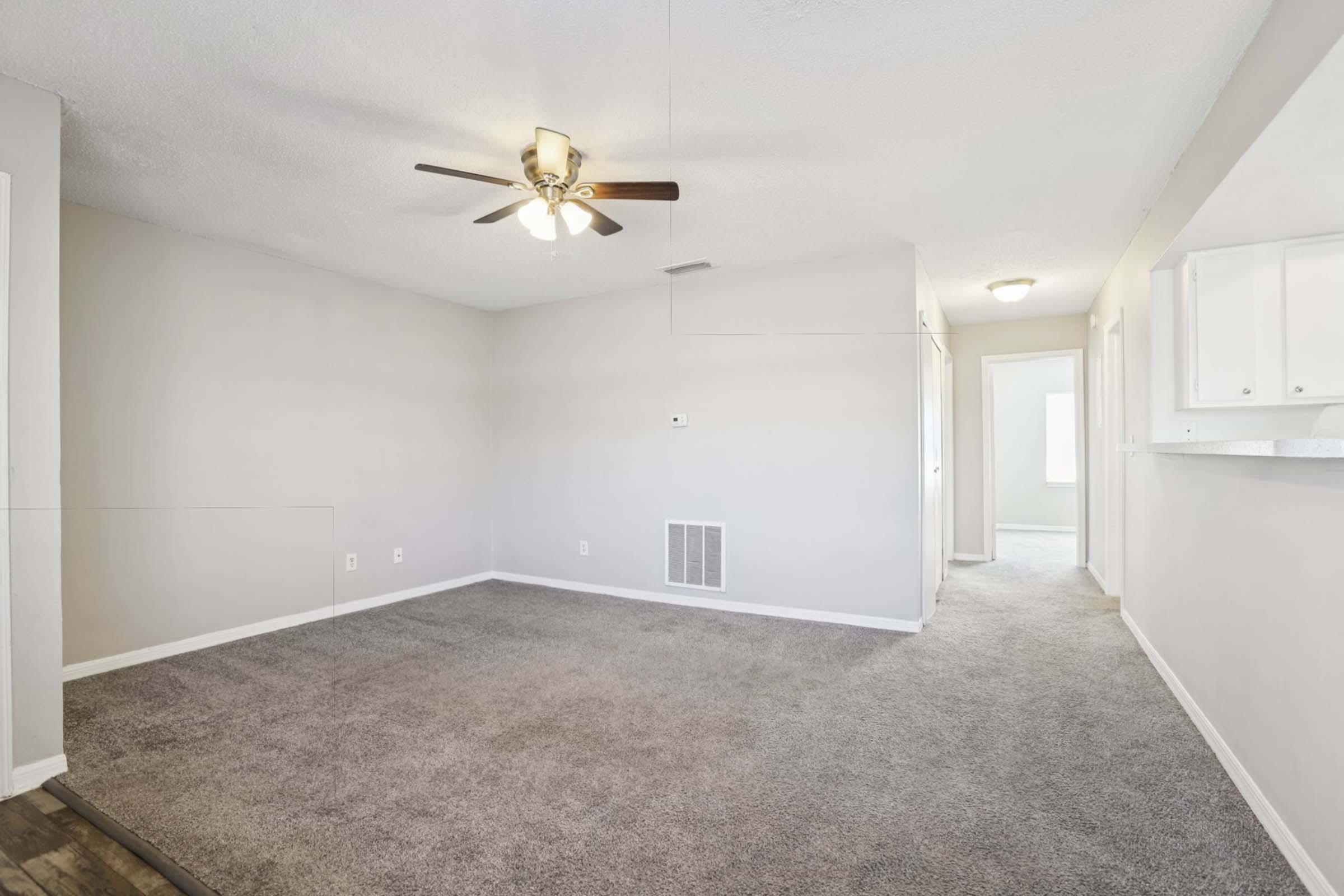 A spacious living room featuring light gray walls, a ceiling fan, and plush carpeting. The room is well-lit with natural light and has a doorway leading to another room. The overall aesthetic is neutral and inviting, perfect for various decor styles.