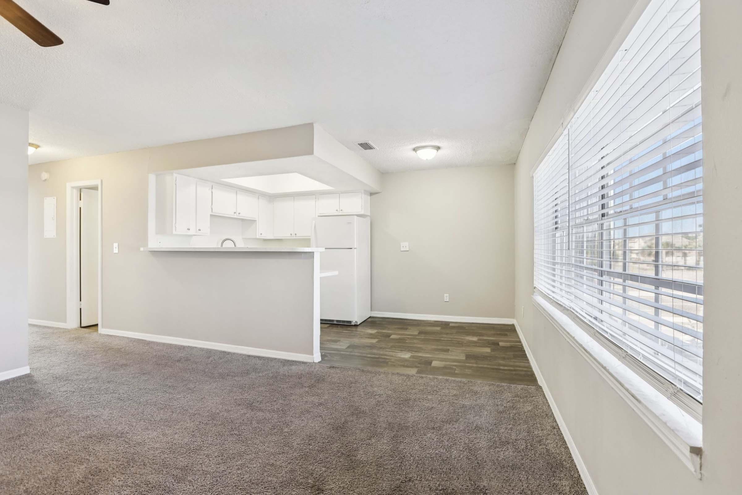 A spacious living area with light-colored walls and carpeted flooring. To the left, there is a kitchen with white cabinets and a refrigerator visible. Large windows in the sunlight allow for natural light, and the overall ambiance is bright and inviting.