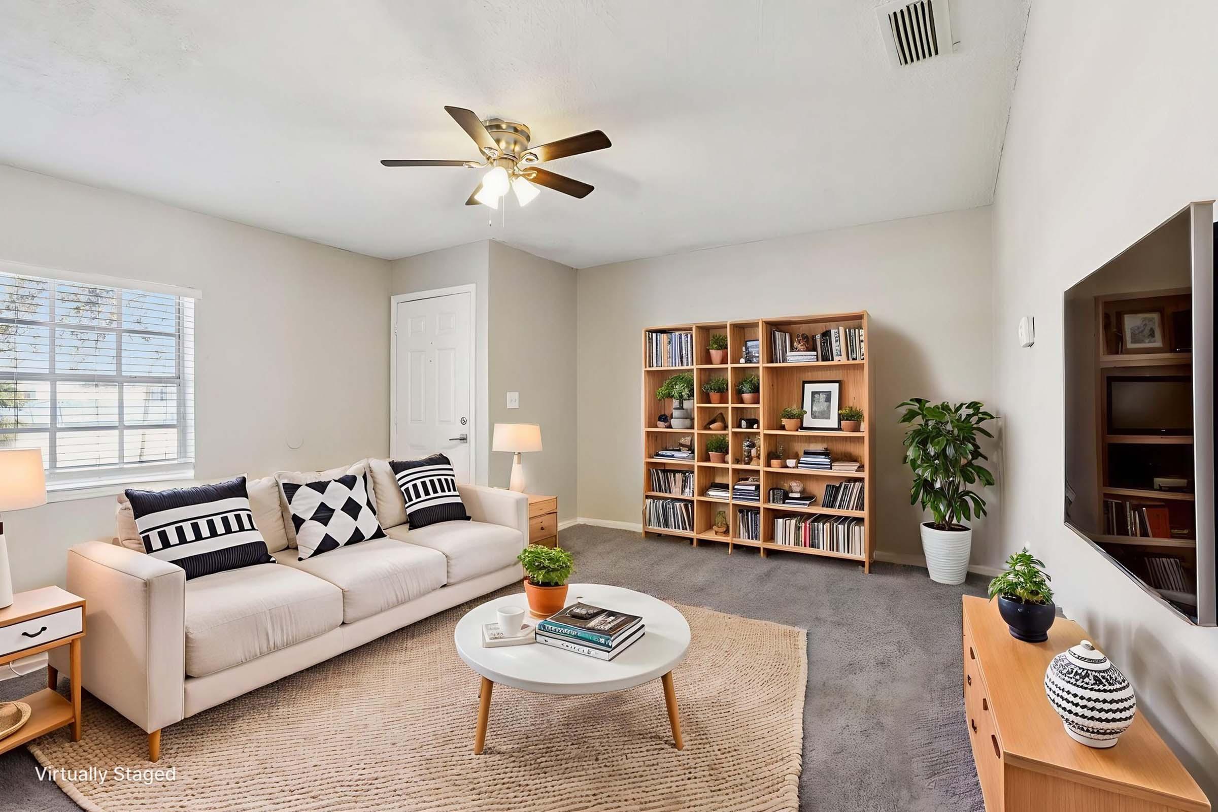 A bright living room featuring a beige sofa with black and white accent pillows, a round coffee table on a textured rug, and a large bookshelf filled with books. A ceiling fan provides lighting, and a potted plant adds greenery. The room has neutral walls and a window letting in natural light, creating an inviting atmosphere.