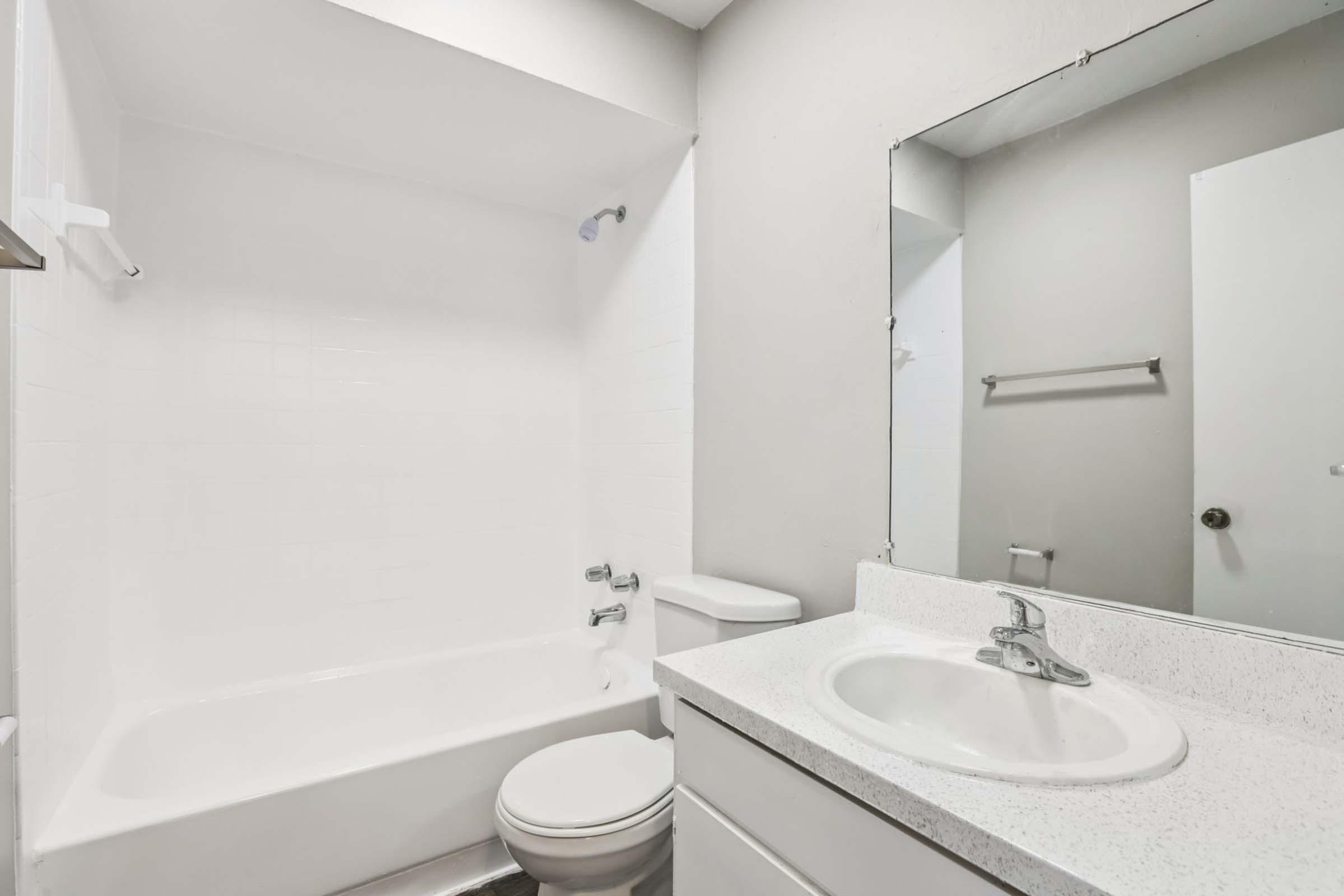 A clean, modern bathroom featuring a white bathtub, toilet, and sink with a countertop. The walls are light-colored, and there is a large mirror above the sink. A small shelf is mounted beside the sink, and the overall design is simple and functional.