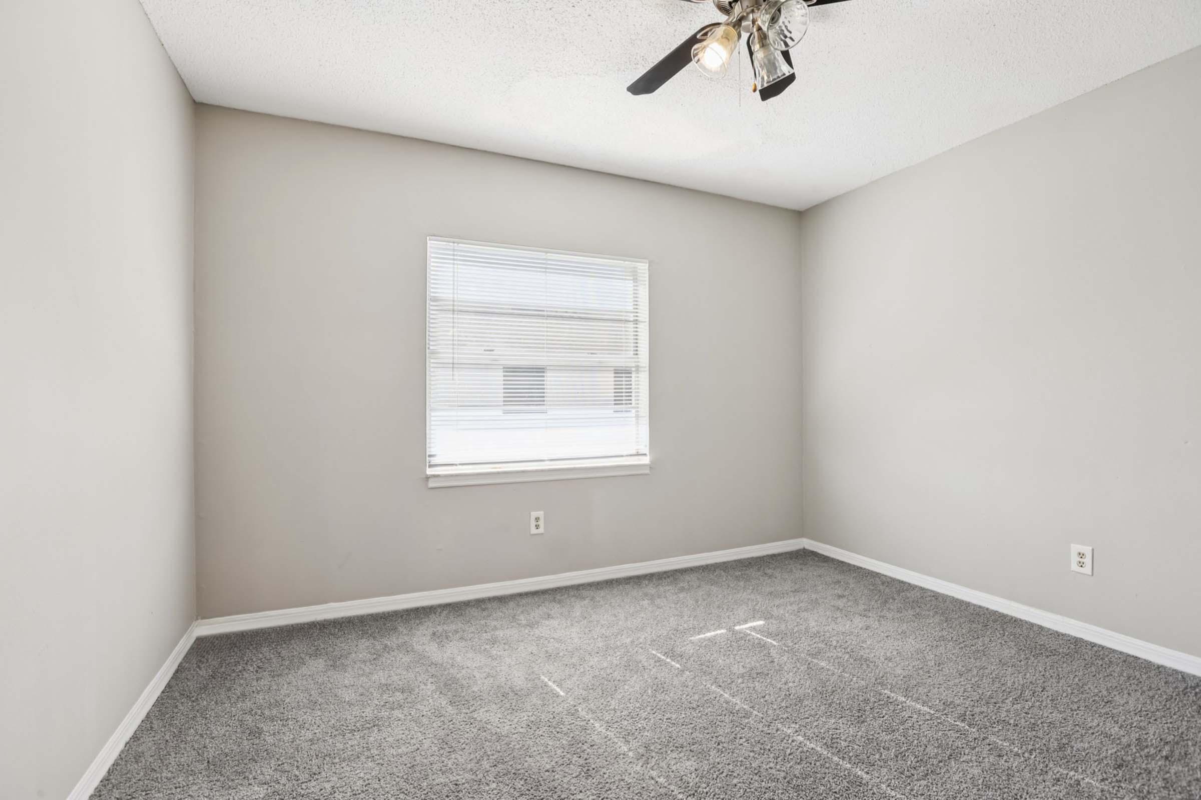 Empty room featuring light gray walls, a ceiling fan, and a window with blinds. The flooring is carpeted in a similar light gray color. There are no furnishings or decorations, creating a clean and minimalist appearance. Natural light enters through the window, illuminating the space.