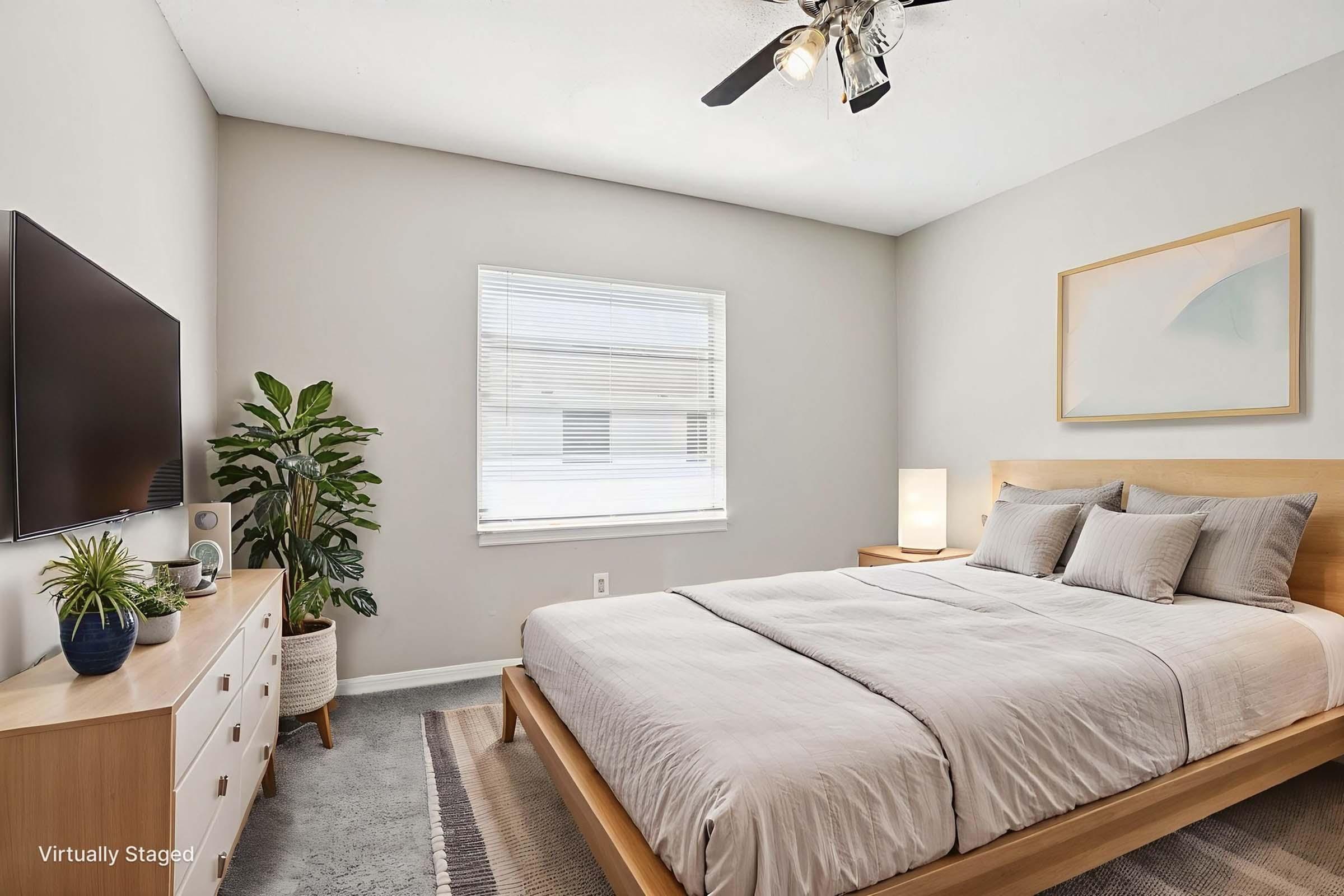 A cozy bedroom featuring a wooden bed frame with a light-colored comforter, two pillows, and a modern nightstand with a lamp. The room has a minimalist design with a potted plant, a TV mounted on the wall, and a window allowing natural light. Soft neutral tones enhance the inviting atmosphere.