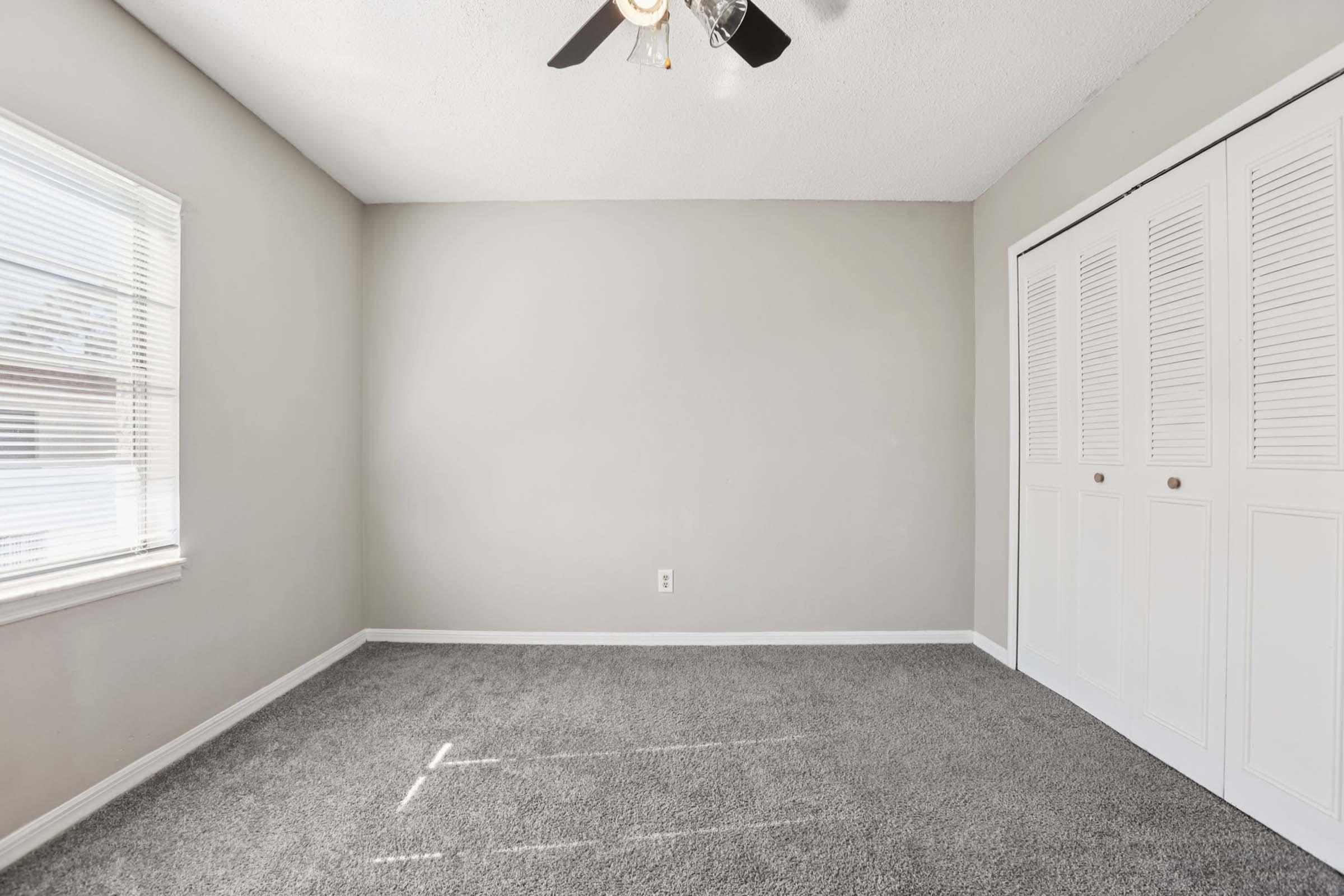 A small, empty room with light gray walls and a ceiling fan. The floor is covered with gray carpet. There is a window with white blinds on the left side, allowing natural light to enter. Opposite the window, there is a white closet with louvered doors. The room appears bright and spacious, suitable for various uses.