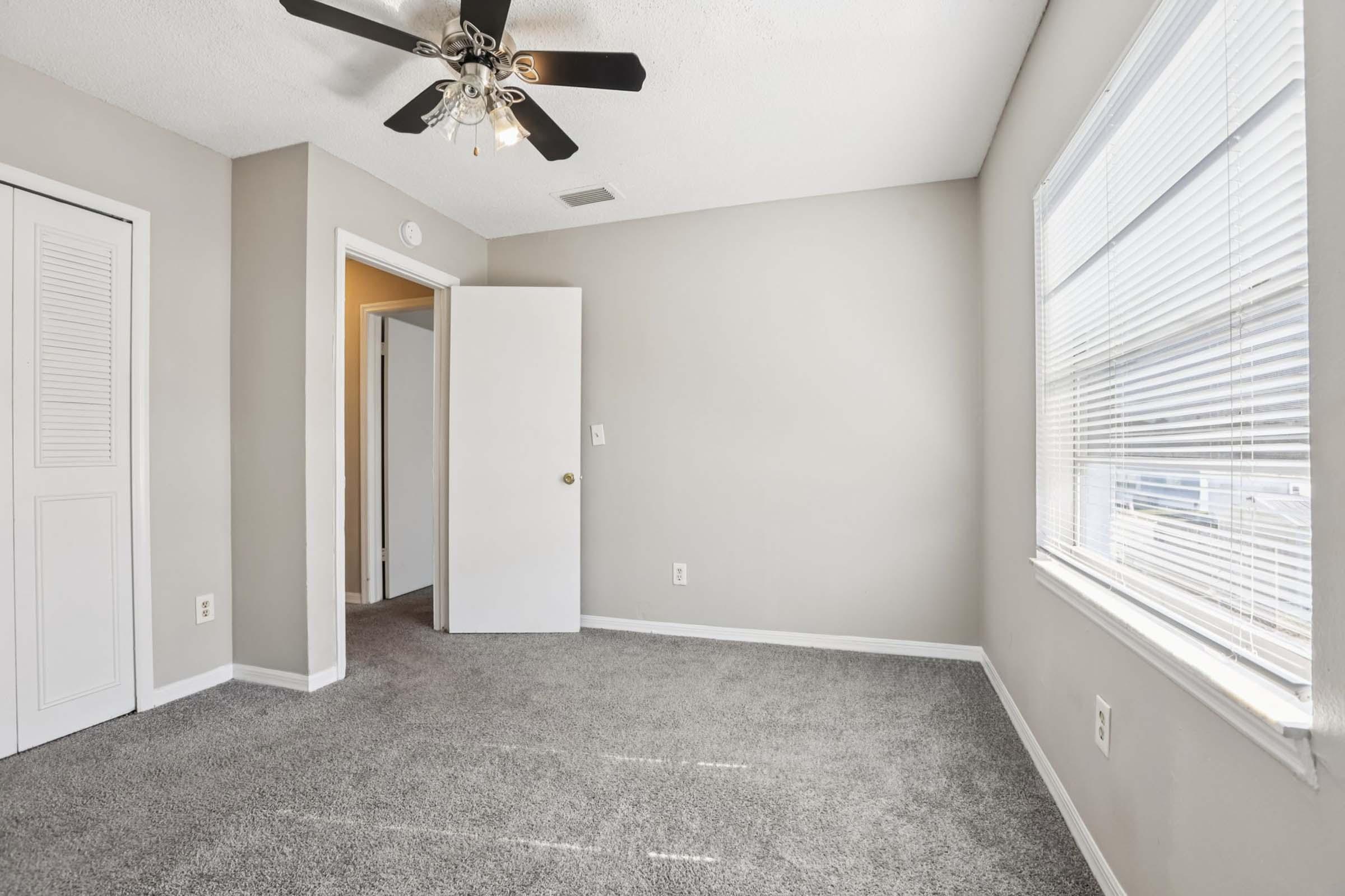A well-lit room with light gray carpet, featuring a ceiling fan, a closet door, and a white door leading to another area. Windows with blinds allow natural light, while the walls are painted in a neutral color. The overall atmosphere is spacious and inviting.