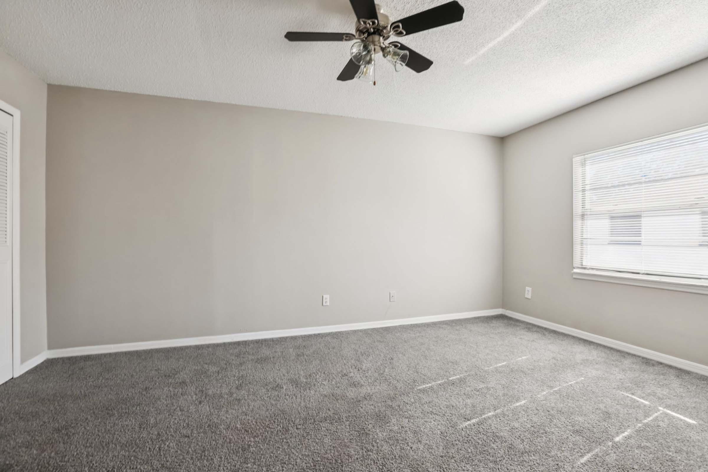 A spacious, empty room featuring light gray walls, a ceiling fan with five blades, and a large window with blinds allowing natural light. The floor is covered in gray carpet, providing a cozy atmosphere. The room is unfurnished, creating a blank canvas for personal decor.