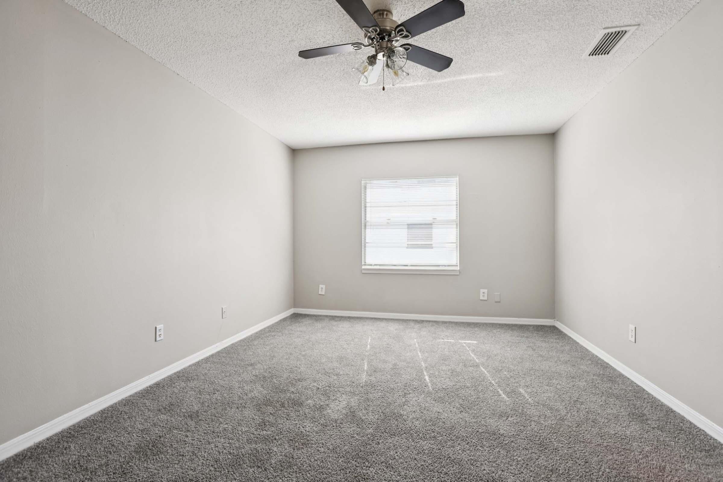 Empty room with light gray walls and plush gray carpet. A ceiling fan with light fixtures is mounted on the ceiling, and there is a single window on one wall allowing natural light to enter. The room has a simple, clean design with no furniture or decorations.