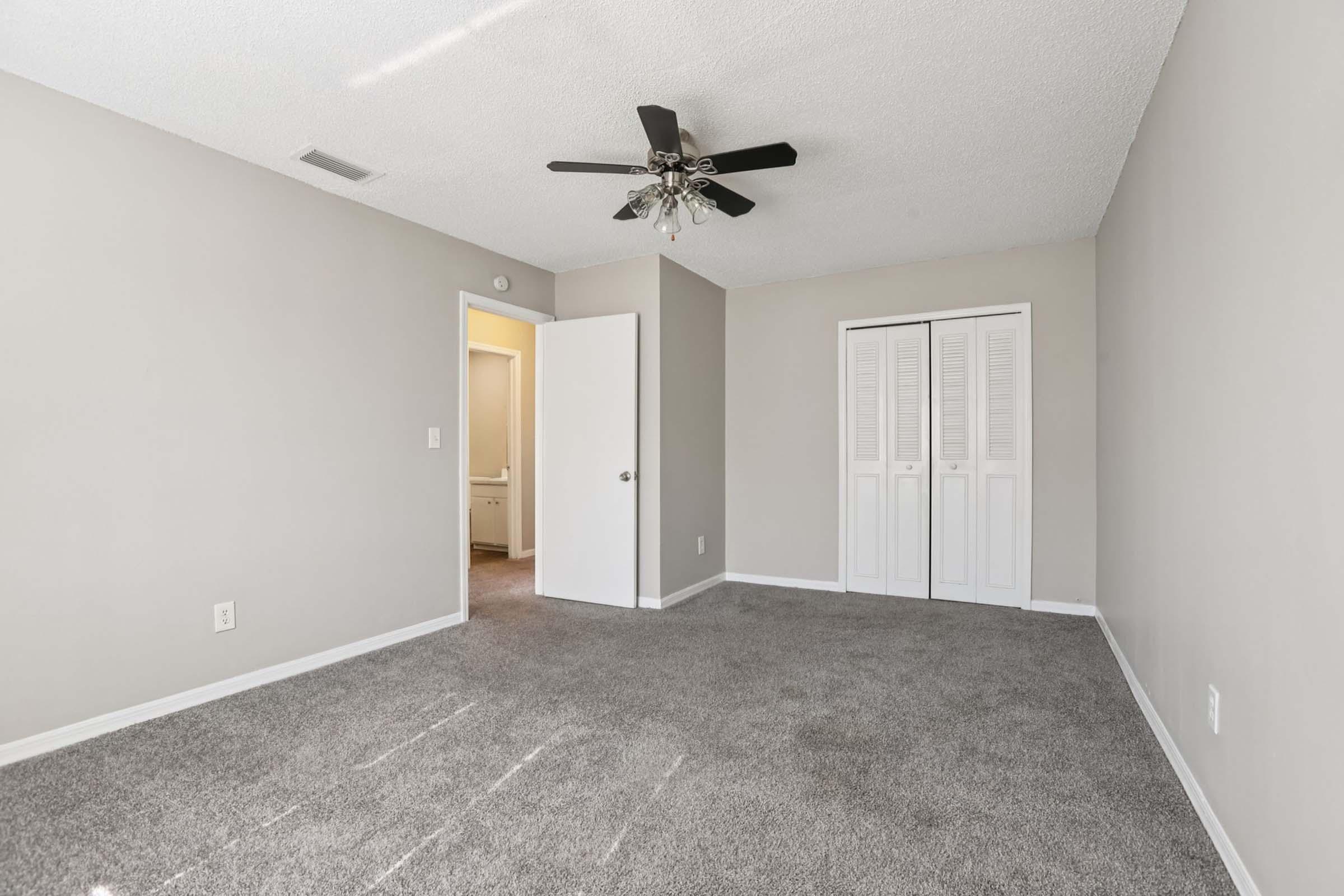 A spacious, empty room with light gray walls and carpet. It features a ceiling fan, a closet with white sliding doors, and a door leading to an adjacent area. Natural light enters from a doorway on the left. The room is ready for decoration and furnishing.
