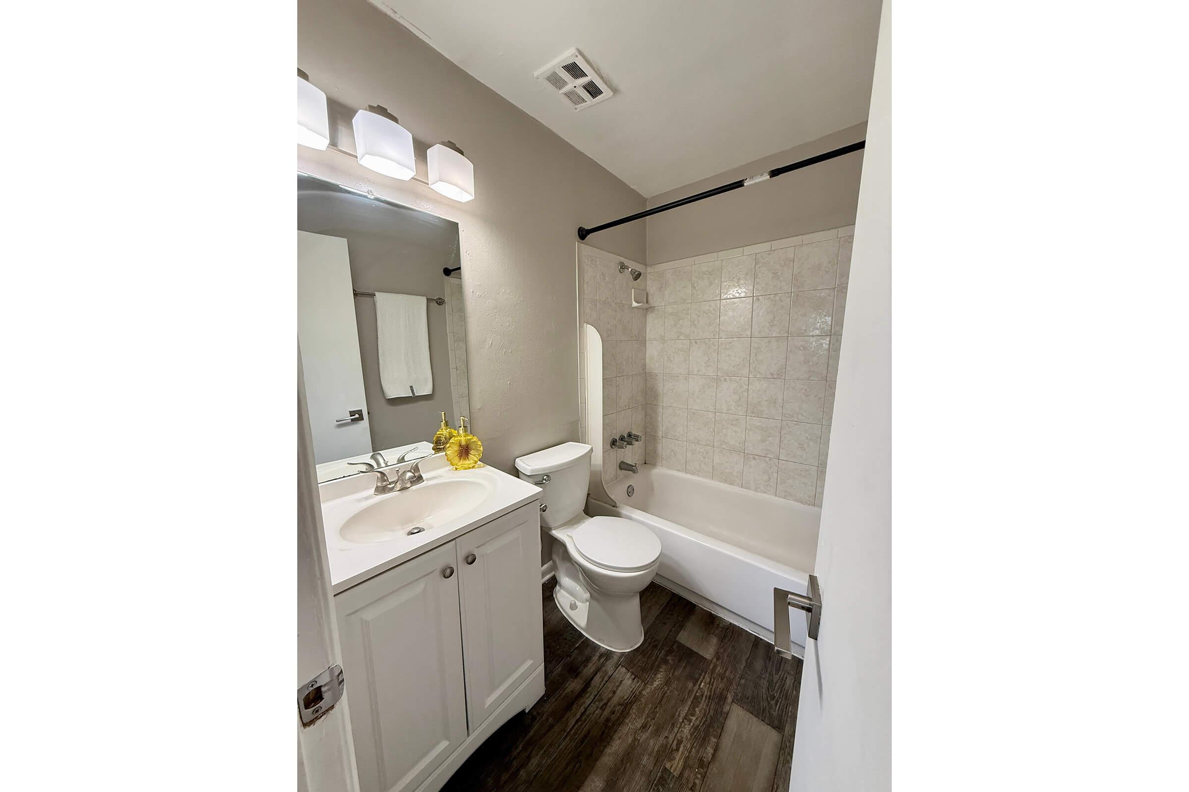 A clean, modern bathroom featuring a white bathtub and shower combination, a toilet, and a sink with a cabinet. The walls are painted light gray, and there is a towel hanging on a rack. Soft lighting is provided by three wall-mounted fixtures, and a small vase of yellow flowers adds a touch of color.
