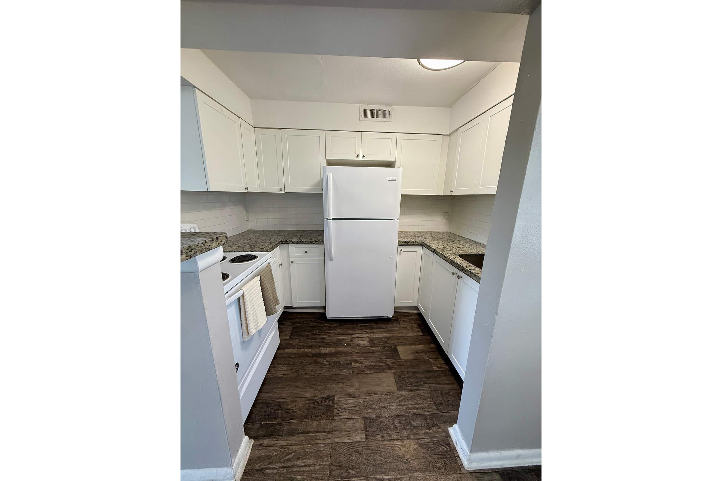 A modern kitchen featuring white cabinetry, a white refrigerator, and a washing machine. The countertops are made of granite, and the floor is adorned with dark wood planks. Soft lighting illuminates the space, creating a bright and clean atmosphere.