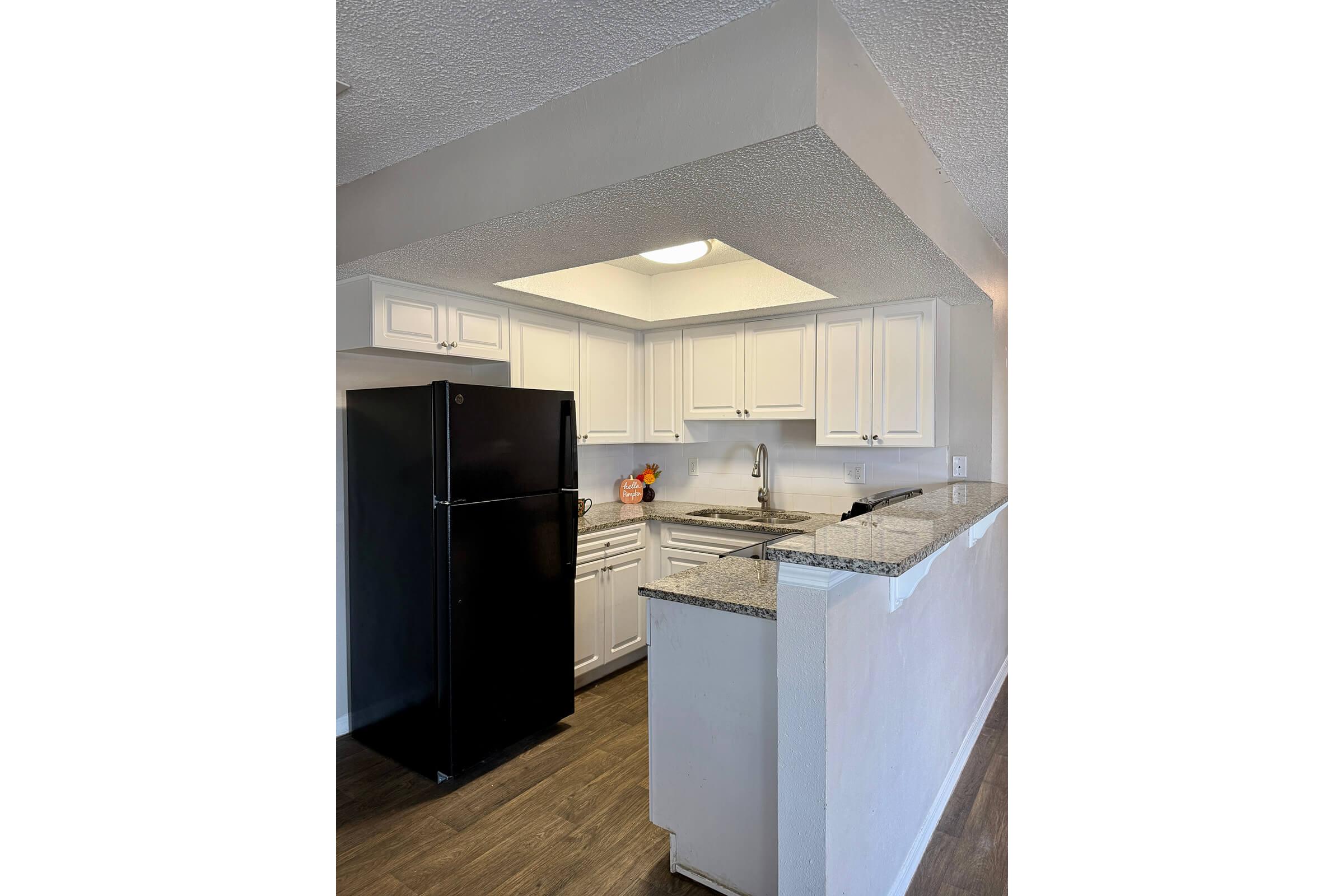 A modern kitchen featuring white cabinetry, a black refrigerator, and granite countertops. The space is well-lit with overhead lighting and includes a sink and dishwasher. The flooring is a warm, wood-like finish, creating a cozy atmosphere.
