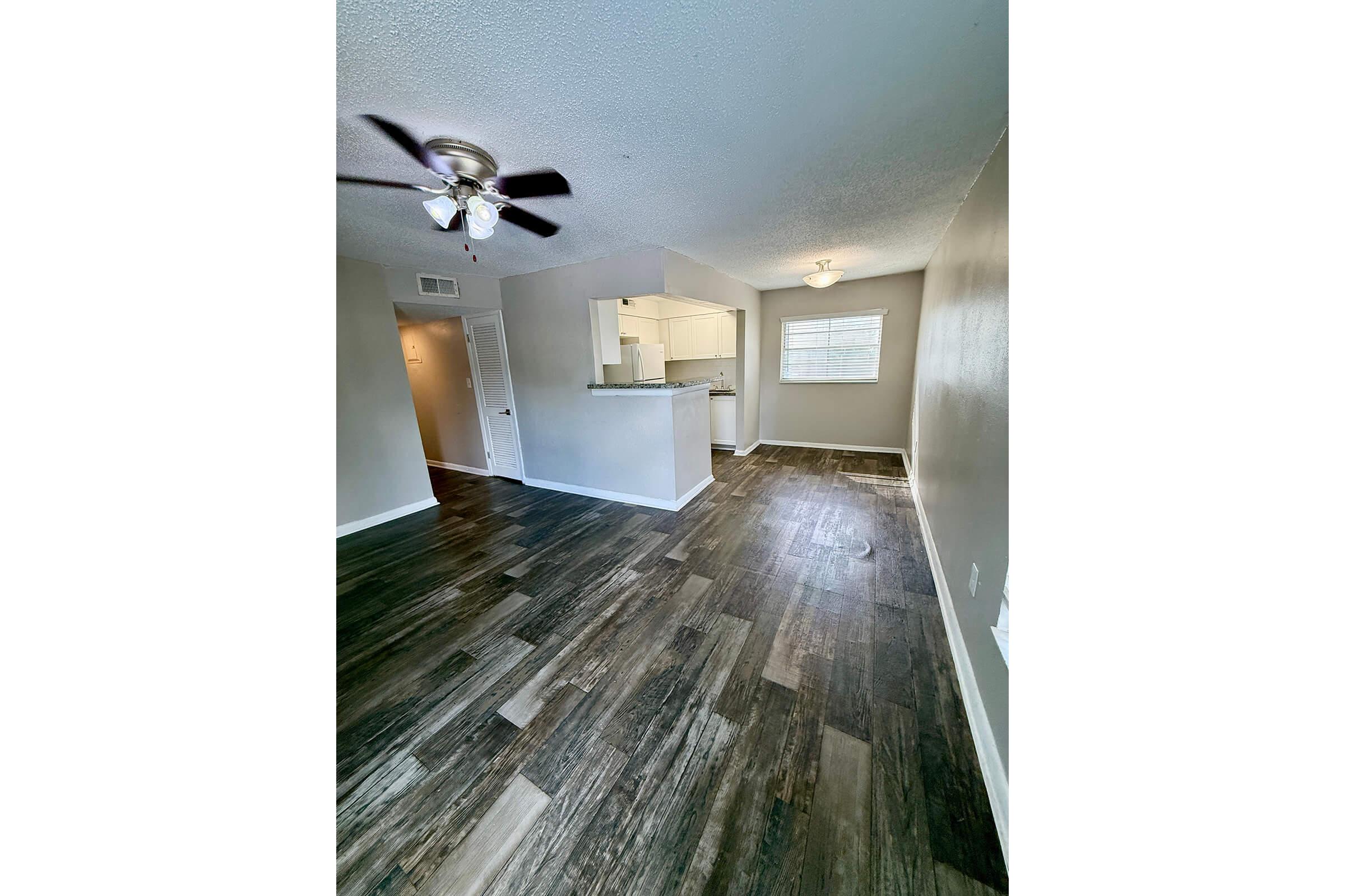 A spacious, well-lit living area featuring dark vinyl flooring, a ceiling fan, and a light-colored wall. In the background, part of a kitchen is visible with white cabinetry. A window lets in natural light, enhancing the inviting atmosphere of the room.