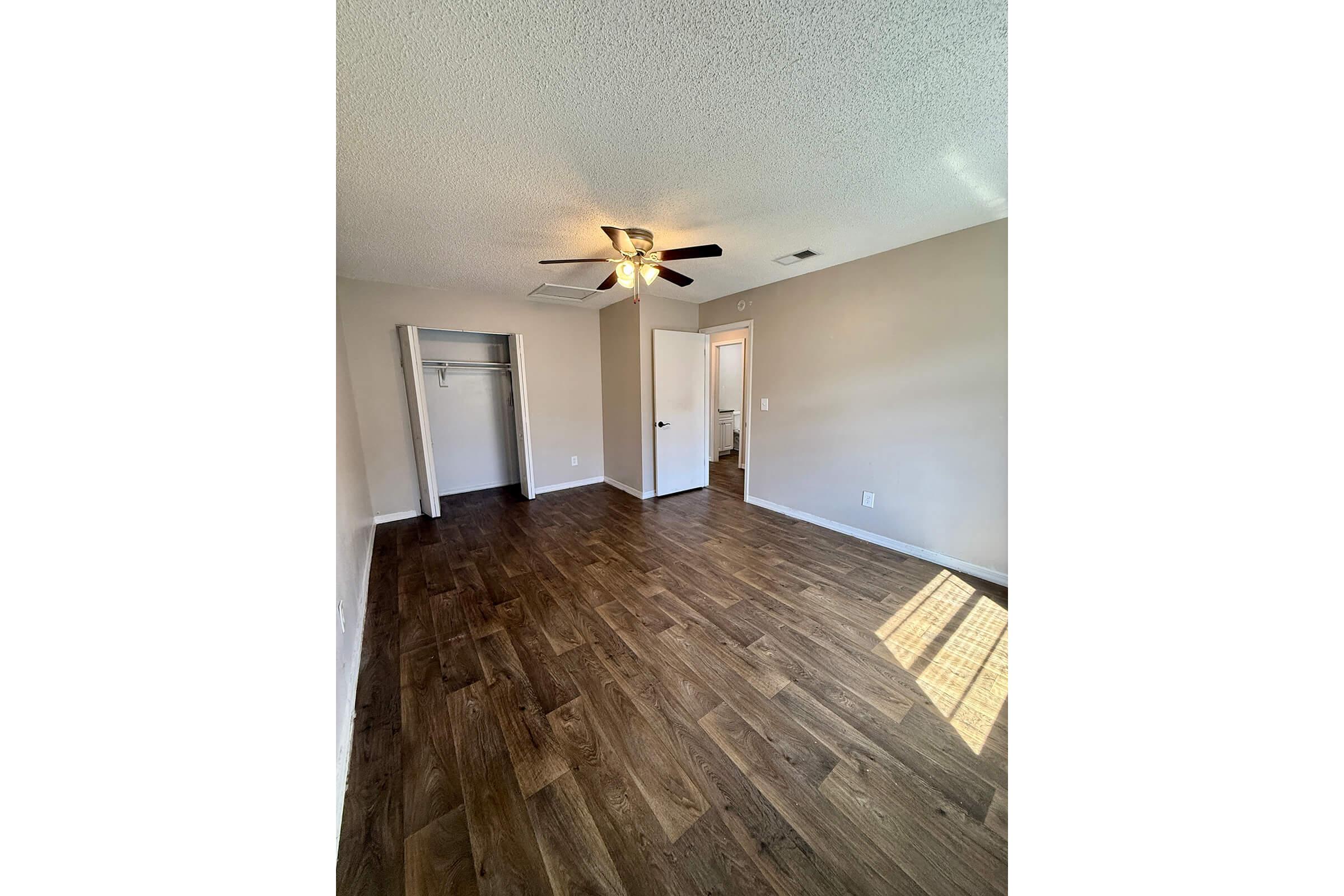 A spacious, empty room with hardwood-style flooring, light beige walls, and a ceiling fan. There is a closet to the left and a doorway leading to another room on the right. Natural light enters through a window, casting a soft glow on the floor.