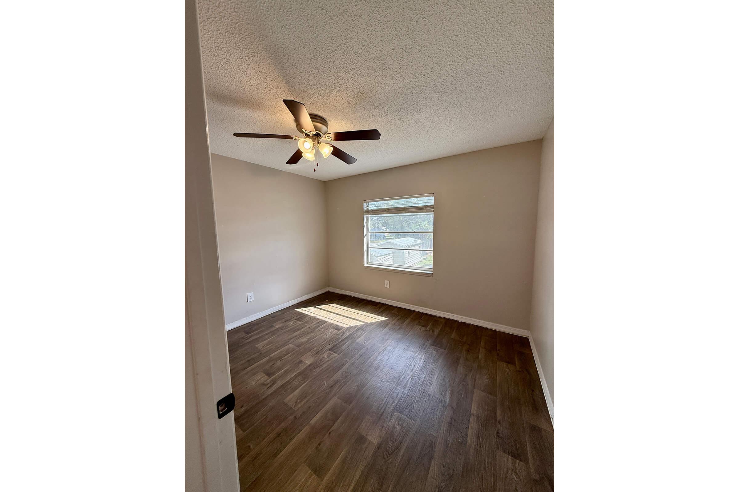 A well-lit, empty room featuring a ceiling fan, a window with light streaming in, and laminate flooring. The walls are painted in a neutral color, creating a spacious and inviting atmosphere.