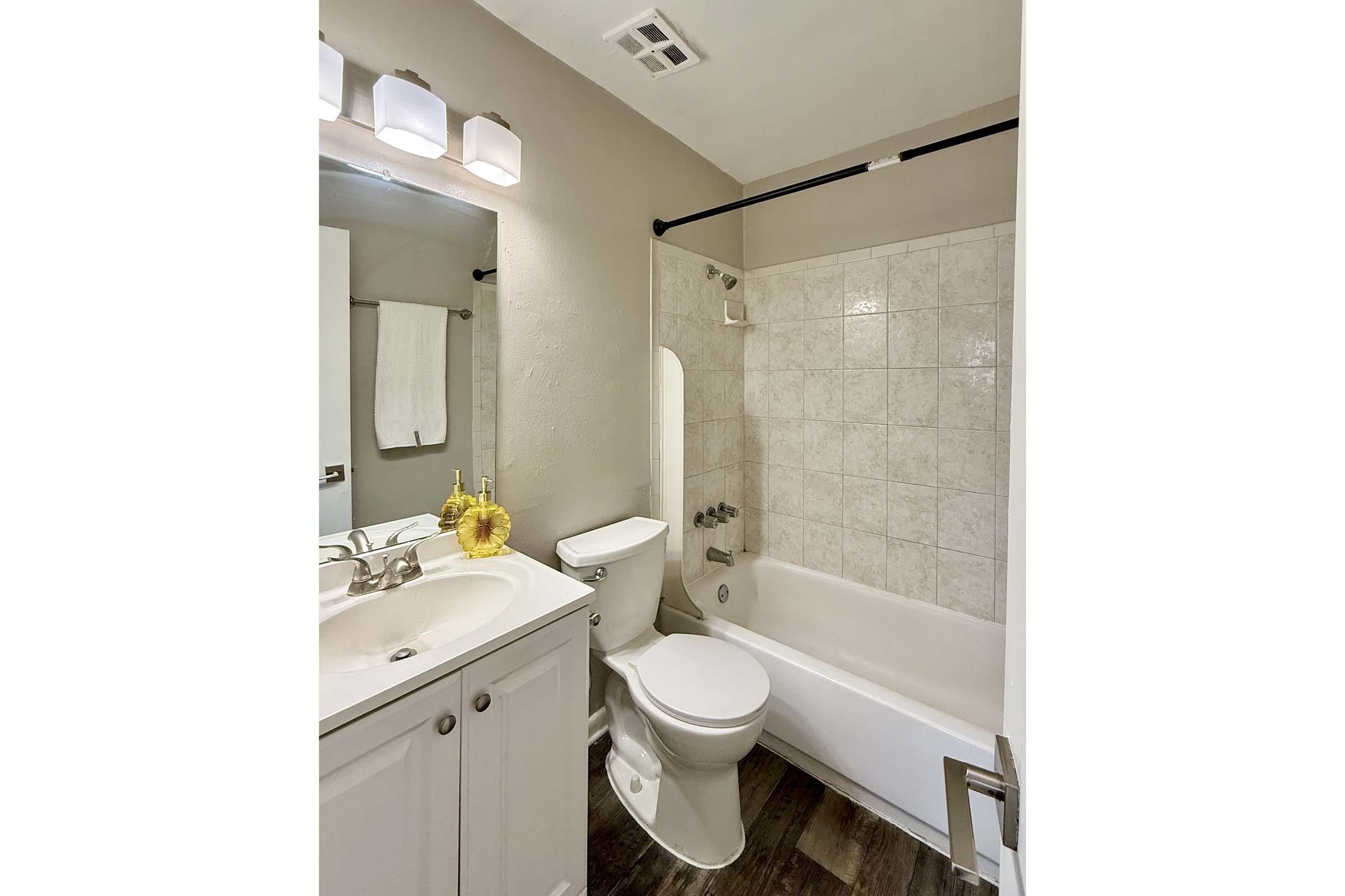 A modern, well-lit bathroom featuring a white sink with a mirror above, a toilet, and a bathtub with a shower. The walls are painted in a neutral color, and there is a shower curtain and a small flower decoration on the counter, creating a clean and inviting atmosphere.
