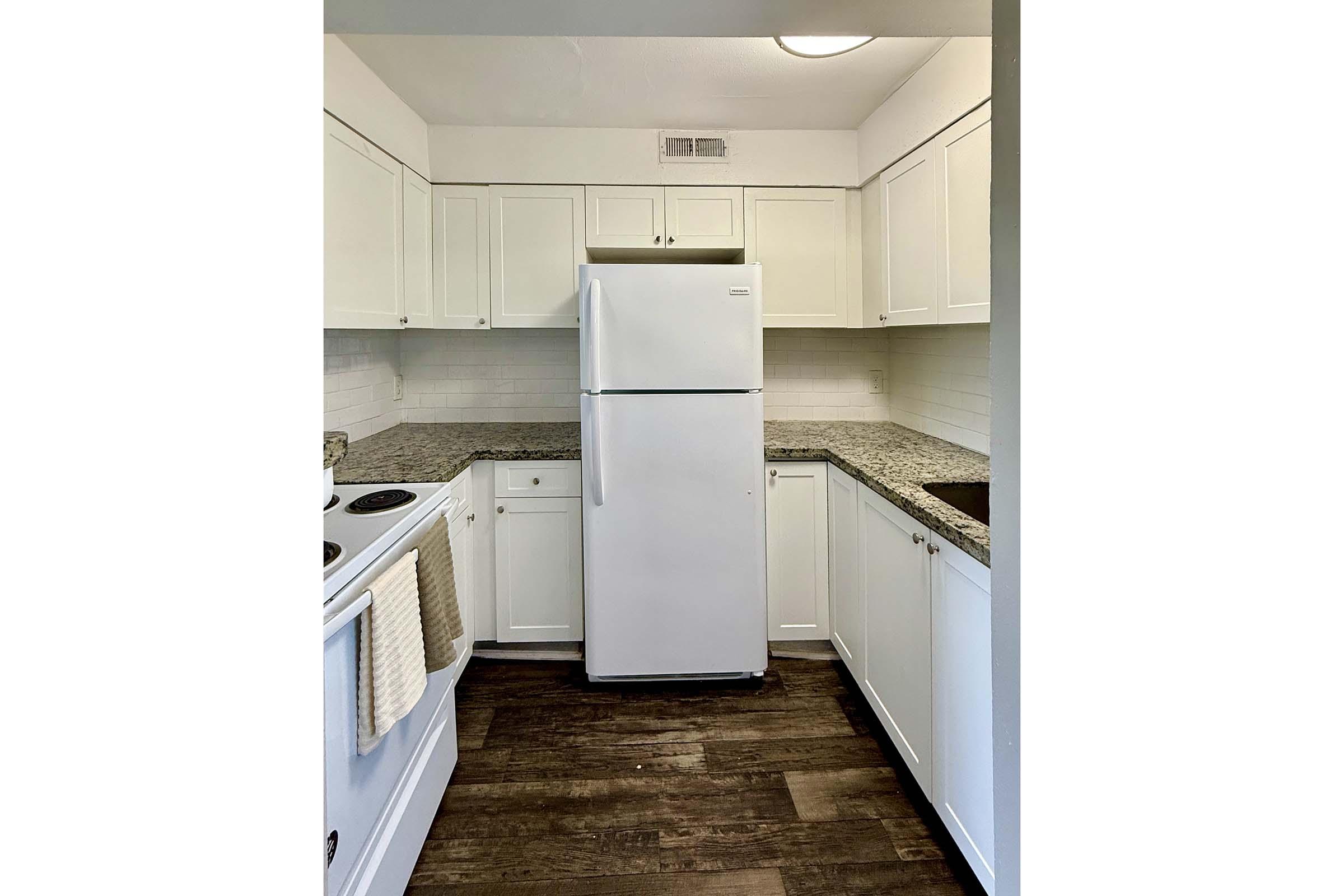 A bright, modern kitchen featuring white cabinetry, a large refrigerator, a stove, and a sink. The countertops are made of granite, and the flooring is a dark wood finish. Soft lighting illuminates the space, creating a clean and inviting atmosphere.