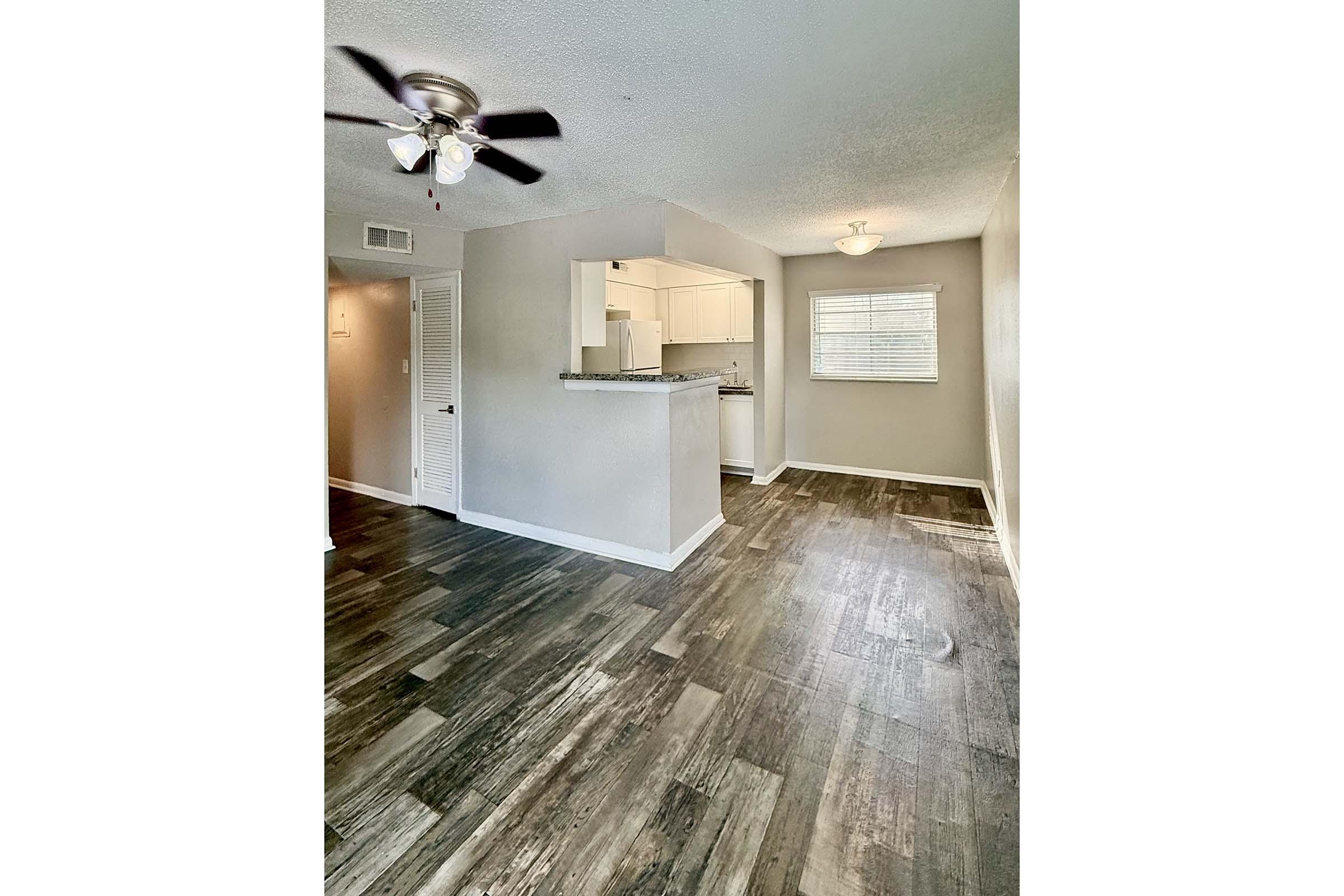 Spacious interior of a modern apartment featuring wood-style flooring, a ceiling fan, and a kitchen visible in the background. Natural light filters in through a window, highlighting the neutral wall colors and open layout, creating a cozy living space.