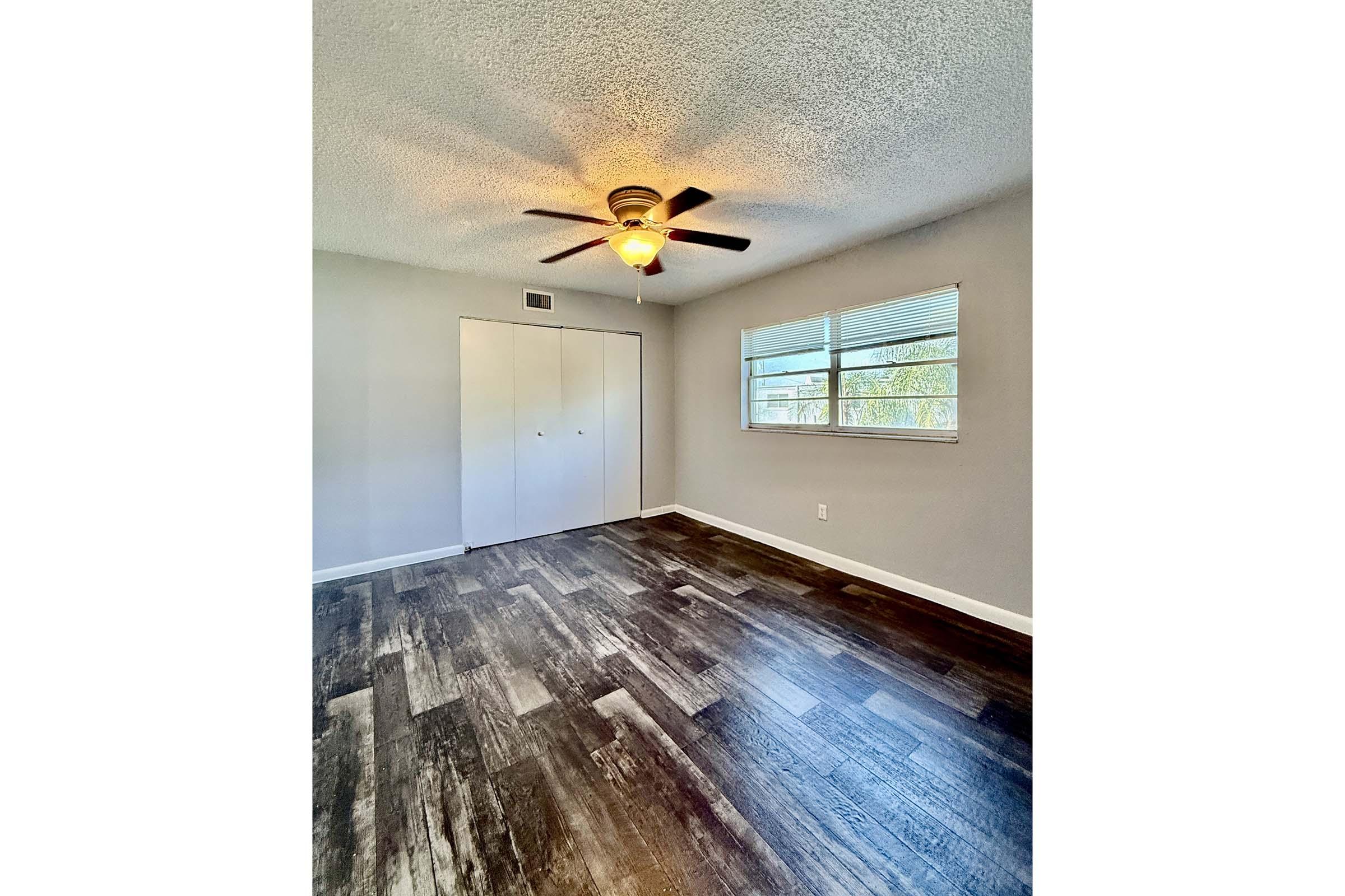 Interior of a room featuring a ceiling fan, large window, and light-colored walls. Dark wooden flooring spans the area, and there are double closet doors on one side, providing ample storage space. The room is well-lit and appears modern and inviting.